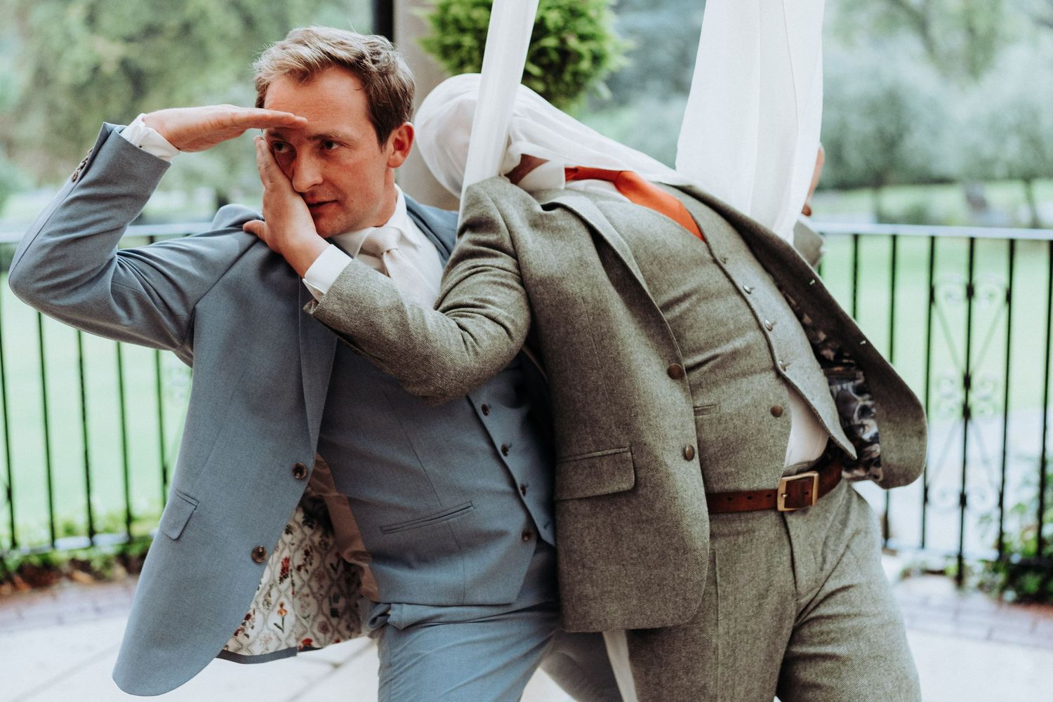 Groom in light grey suit playfully posing with jacket over shoulder at outdoor wedding venue.