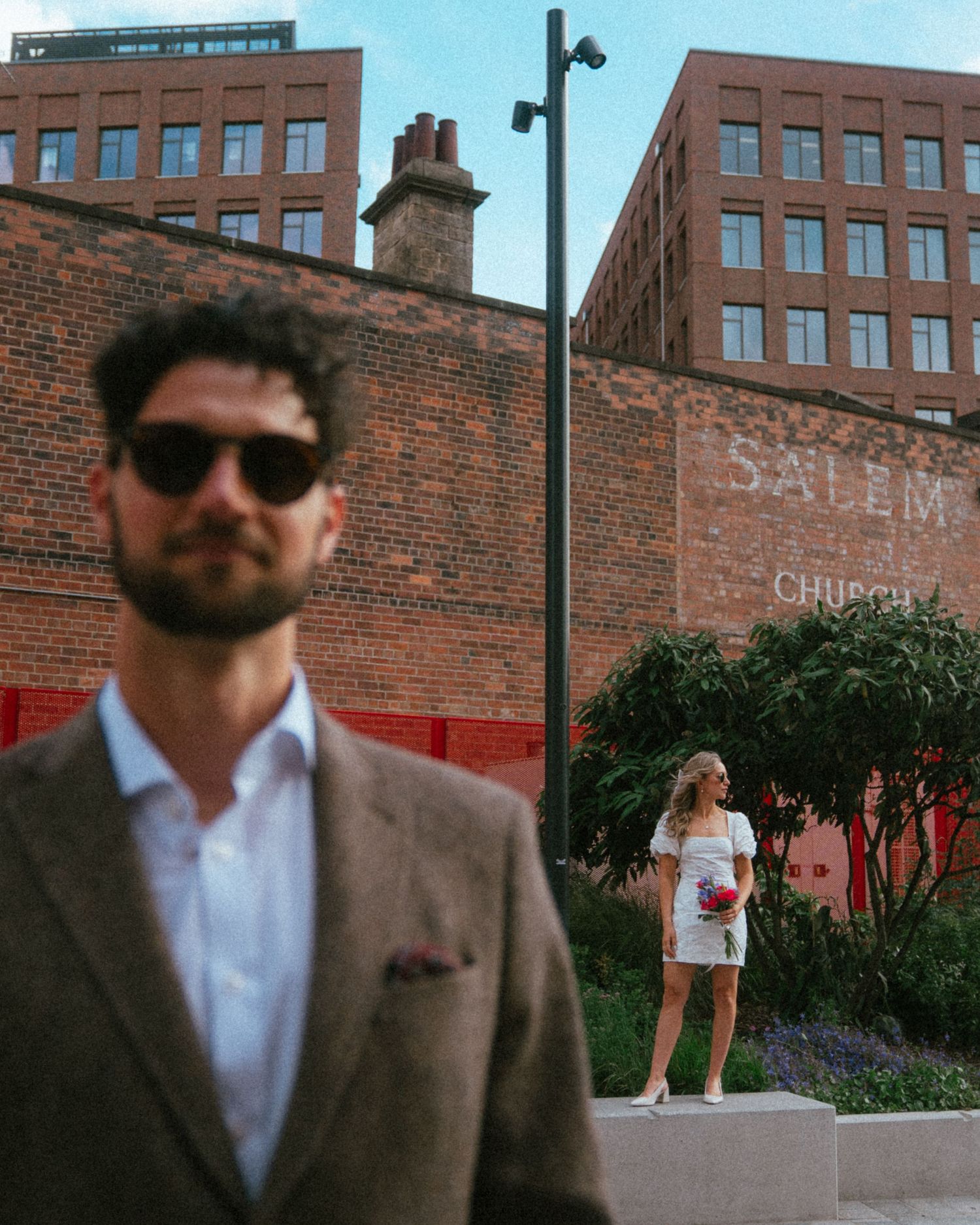 Man in sunglasses and brown blazer with woman in white dress posing near brick buildings with Salem Church sign.