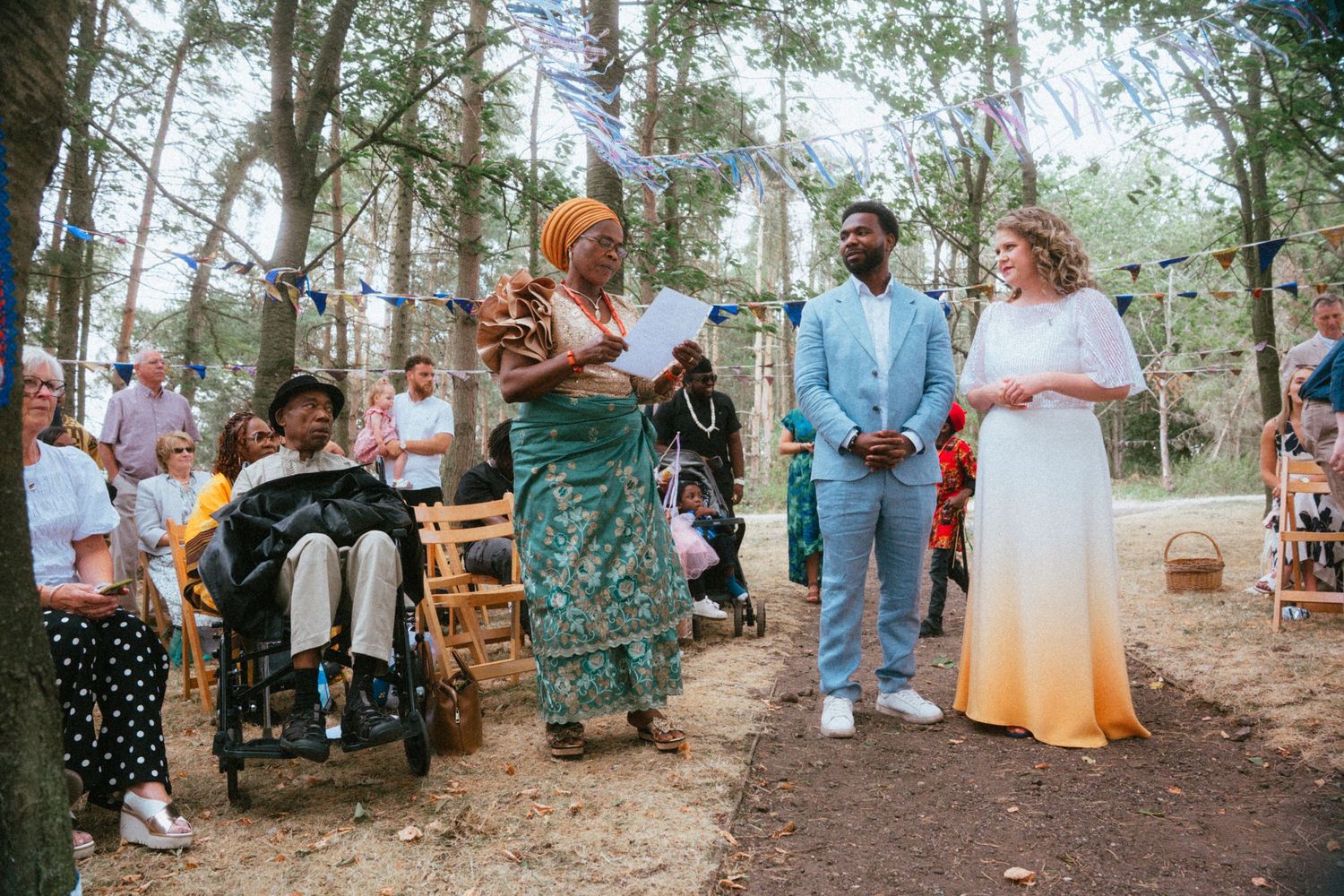 Outdoor woodland wedding ceremony with couple in colorful attire, officiant in teal dress, and guests seated among trees.
