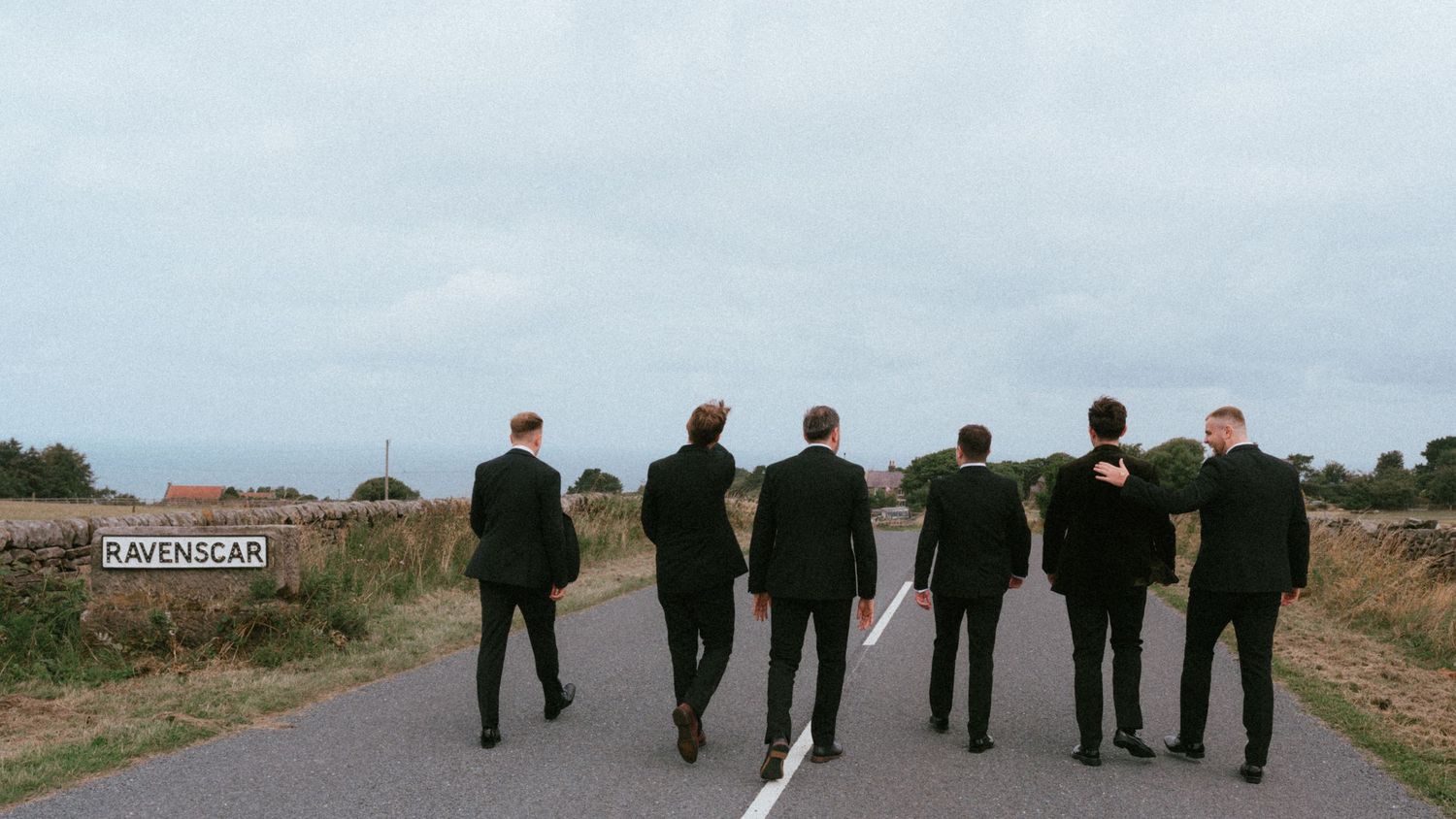 Six men in dark suits walk away from the camera down a rural road toward a Ravenscar village sign under an overcast sky.