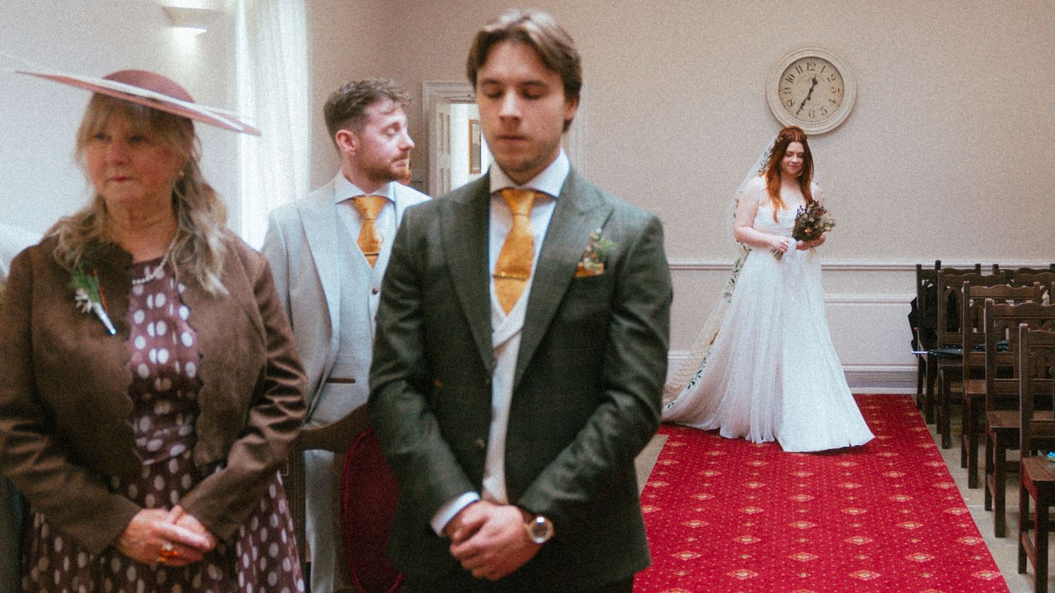 Wedding ceremony scene with bride in white gown on red carpet aisle, groom in gray suit, guests in formal attire.