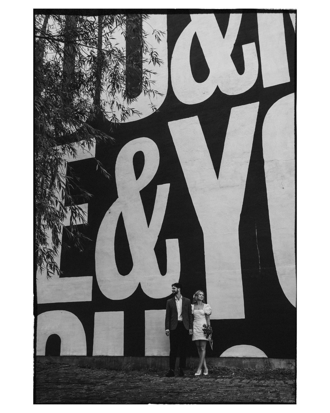 Black and white photo of a couple standing before a large typographic mural with ampersands and bold letters.