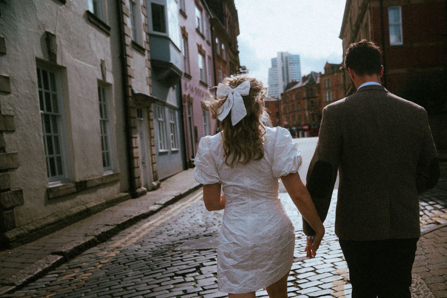 Bride in white mini dress with bow and groom walk hand in hand down a cobblestone street.