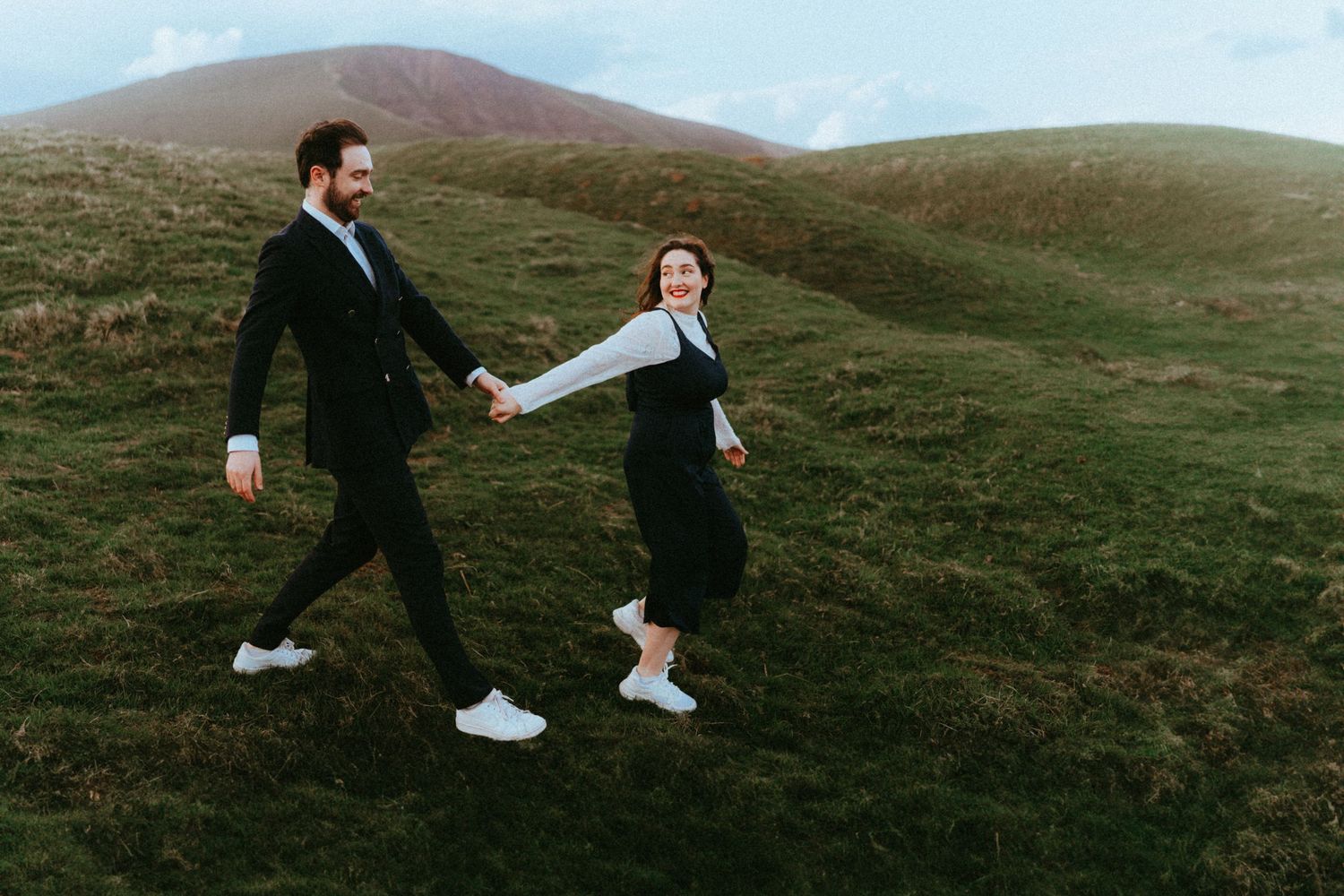 Couple holding hands and walking through lush green highland hills under a dramatic cloudy sky.