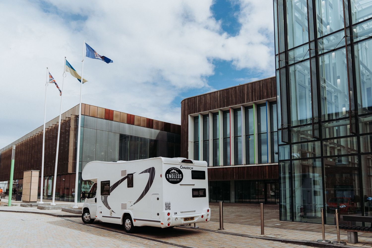 White motorhome parked outside a modern glass building with flags flying on a cloudy day.