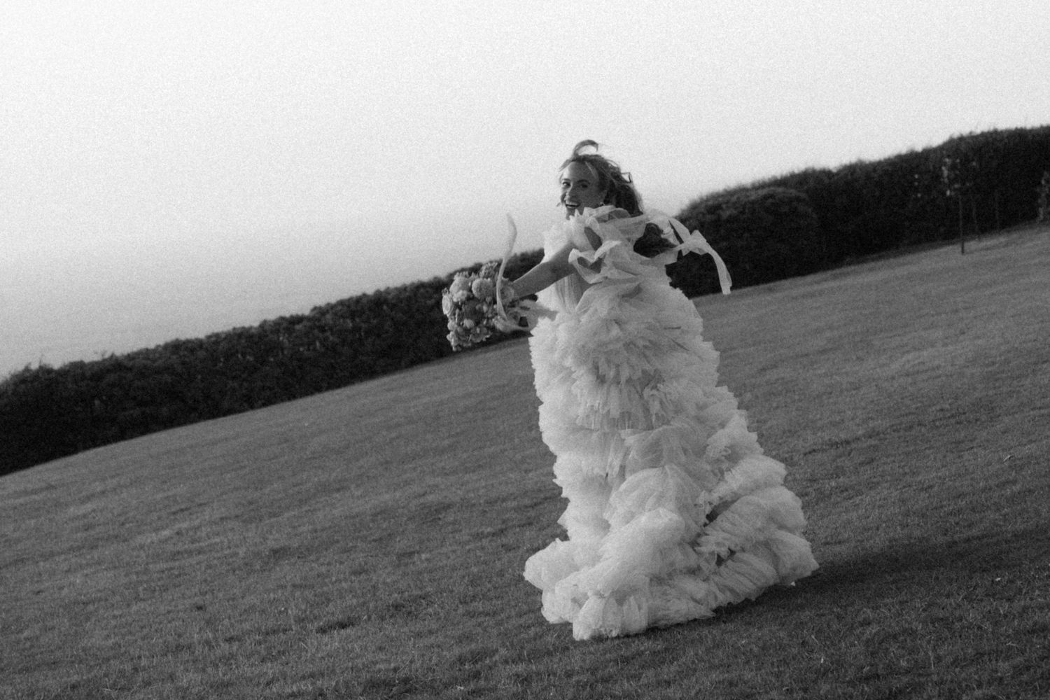 Black and white photo of a bride in a ruffled tulle wedding gown holding a bouquet while twirling on a grassy hillside near a hedge.