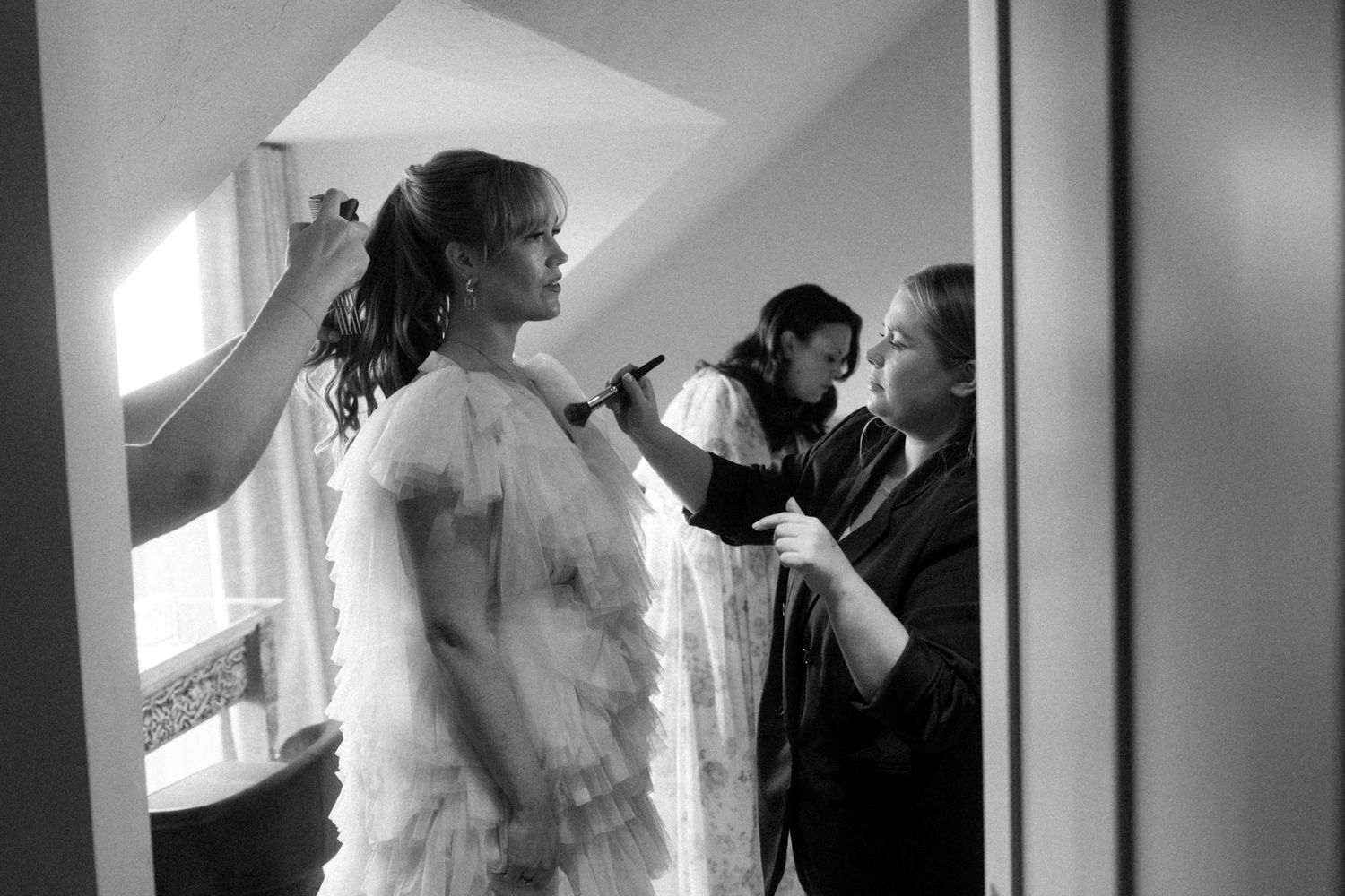 Black and white photo of a bride in a ruffled tulle gown having her hair and makeup done by two stylists in a softly lit room.