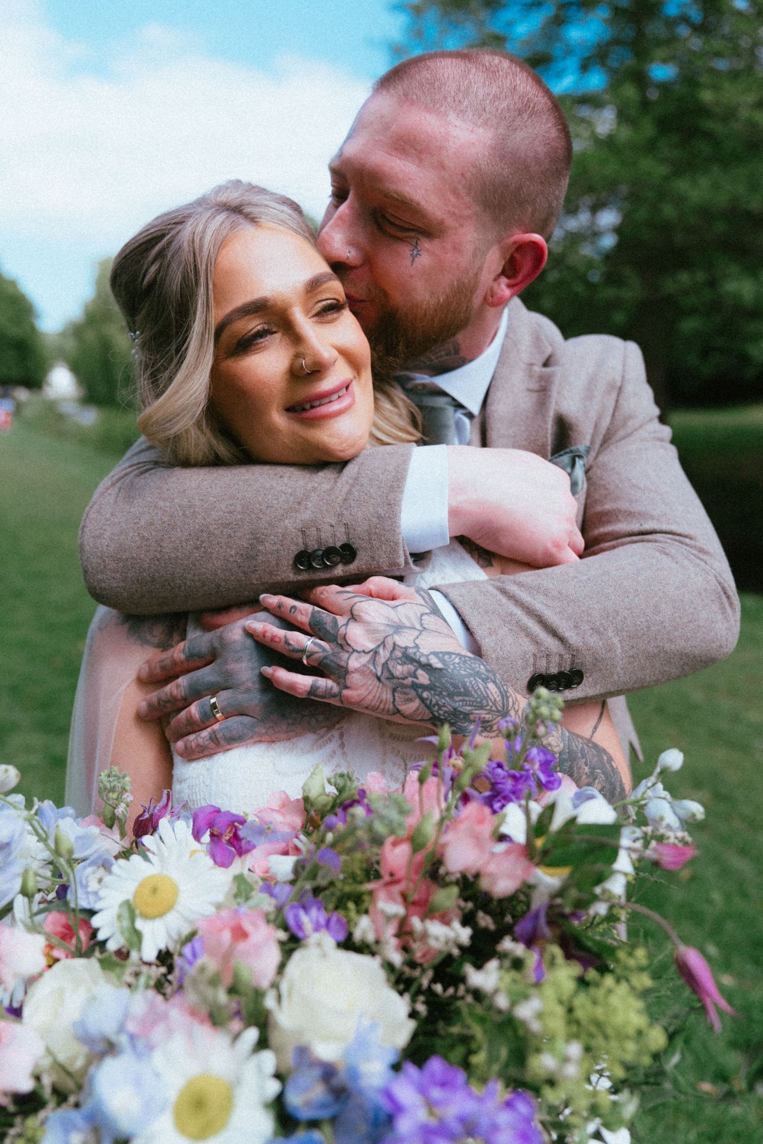 Tattooed groom tenderly kissing bride's cheek while embracing her with floral bouquet below.