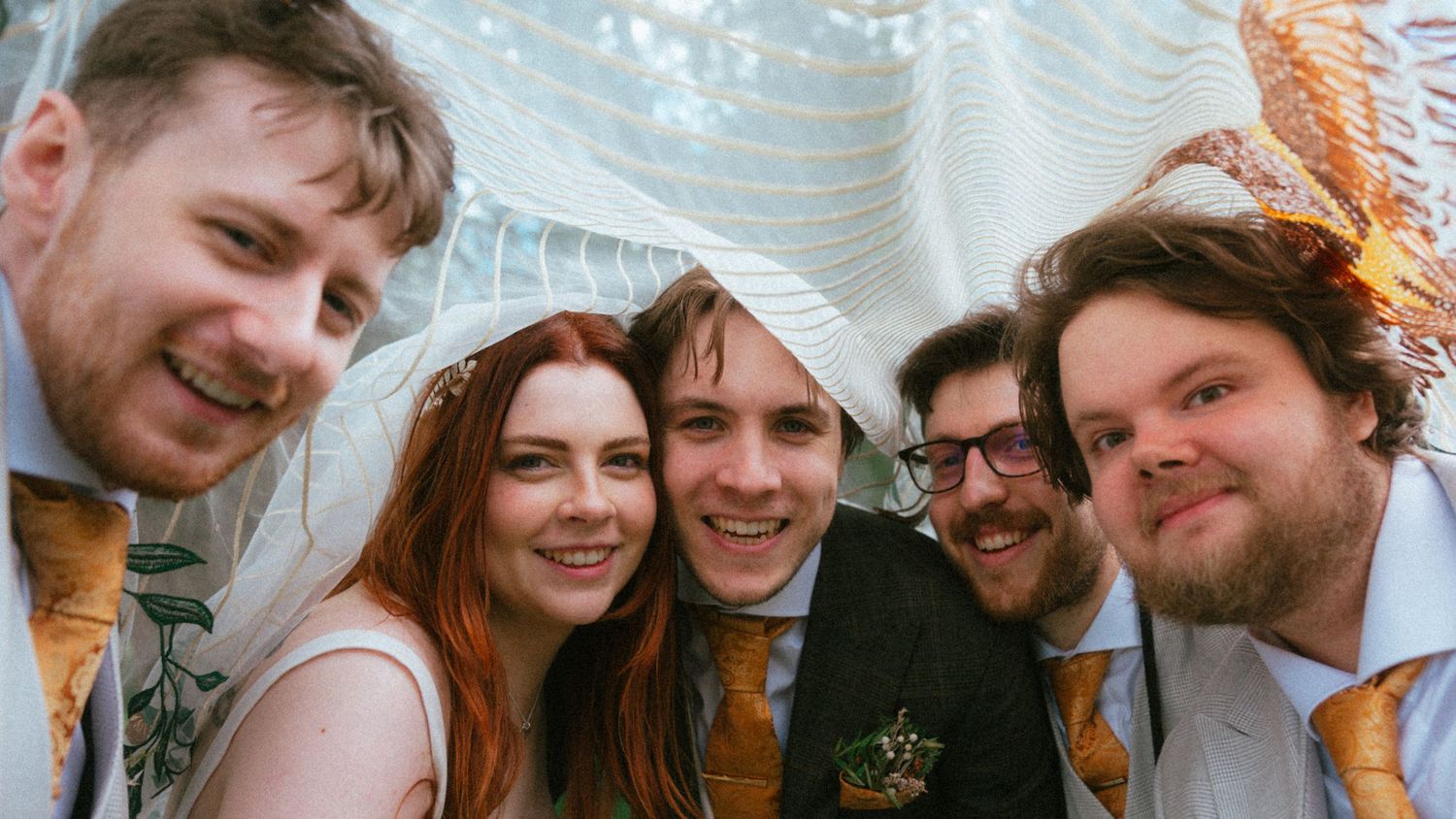 Five smiling friends pose closely together at a wedding, wearing formal attire with gold ties and a white dress.