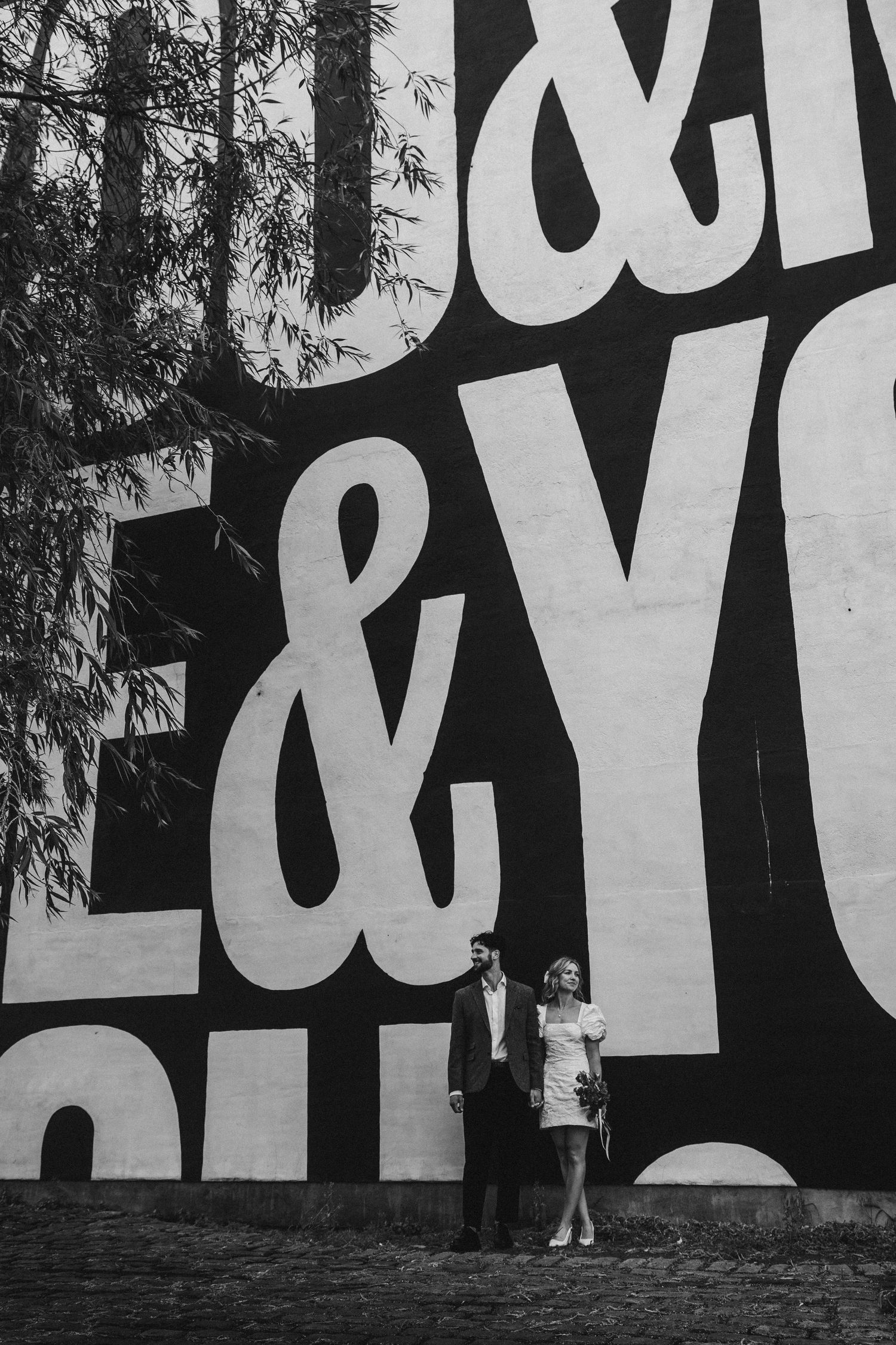 Black and white photo of a couple standing in front of a large mural with bold ampersand typography.