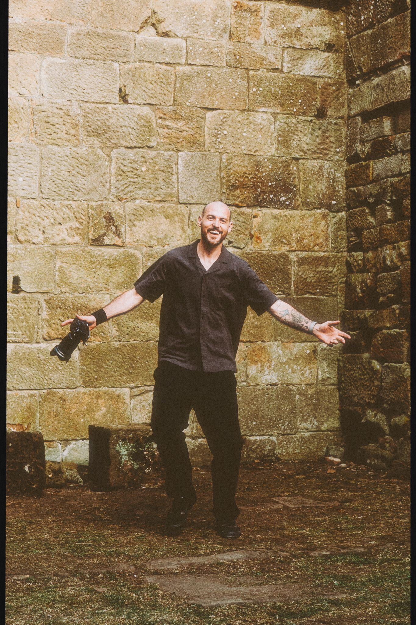 Smiling man with tattoos posing with open arms against a rustic stone wall outdoors.