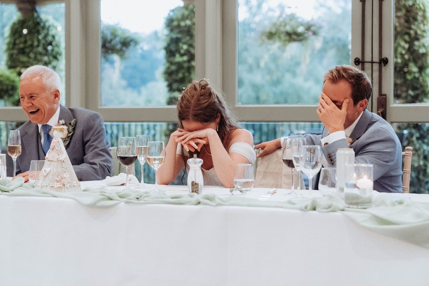 Bride, groom, and guest laugh and cover faces during amusing wedding speech at reception table.