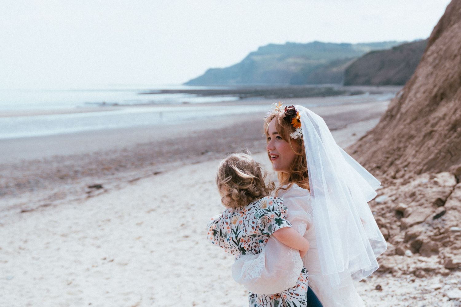 A bride in a white veil and floral hair crown holds a young girl in a floral dress on a sandy beach with cliffs and calm sea behind them.