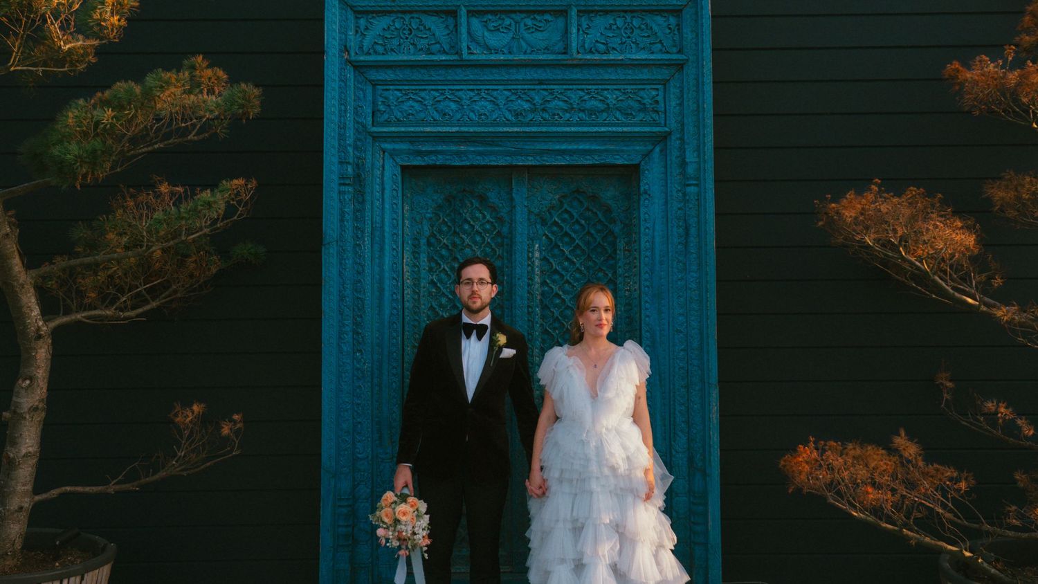 A bride in a ruffled white tulle gown and a groom in a black tuxedo with bow tie stand before a vivid teal door holding a bouquet.