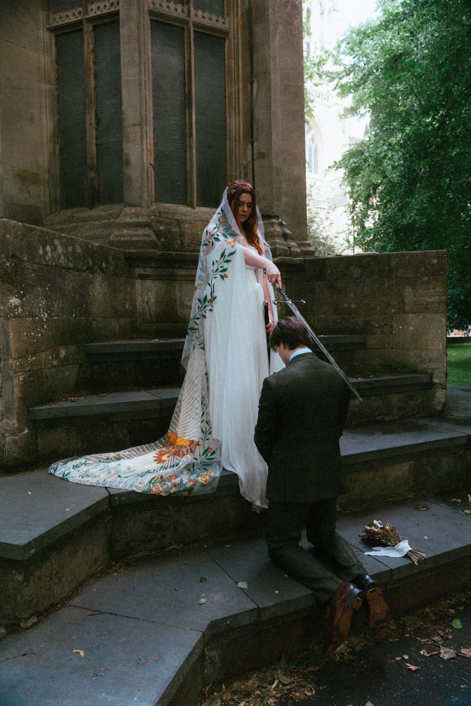 Bride in embroidered veil standing on stone steps while groom kneels beside Gothic church.