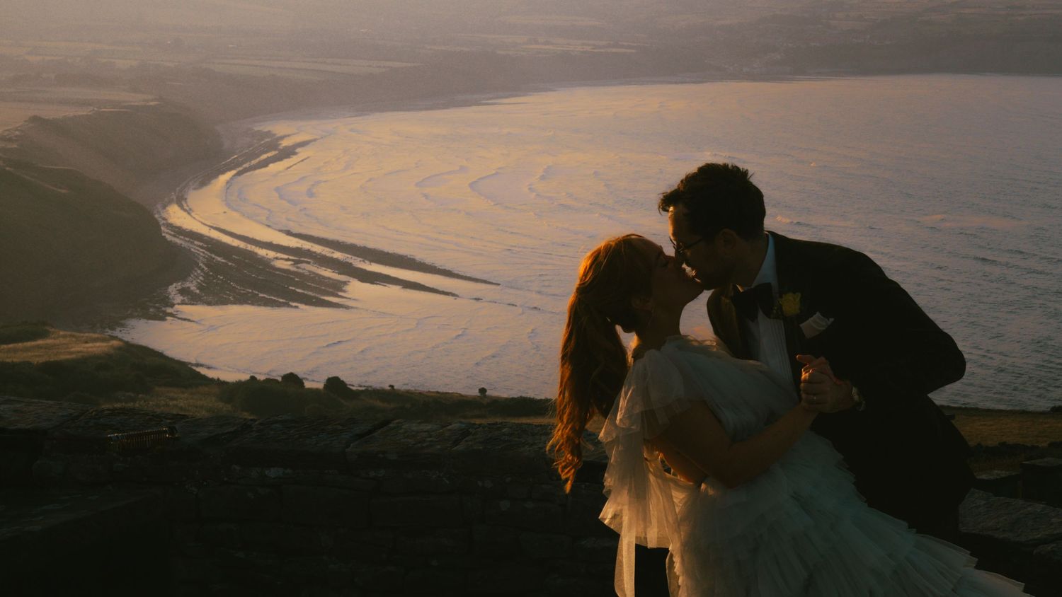 Bride in a white gown and groom in a dark suit share a romantic kiss on a hilltop overlooking a golden sunset coastline.