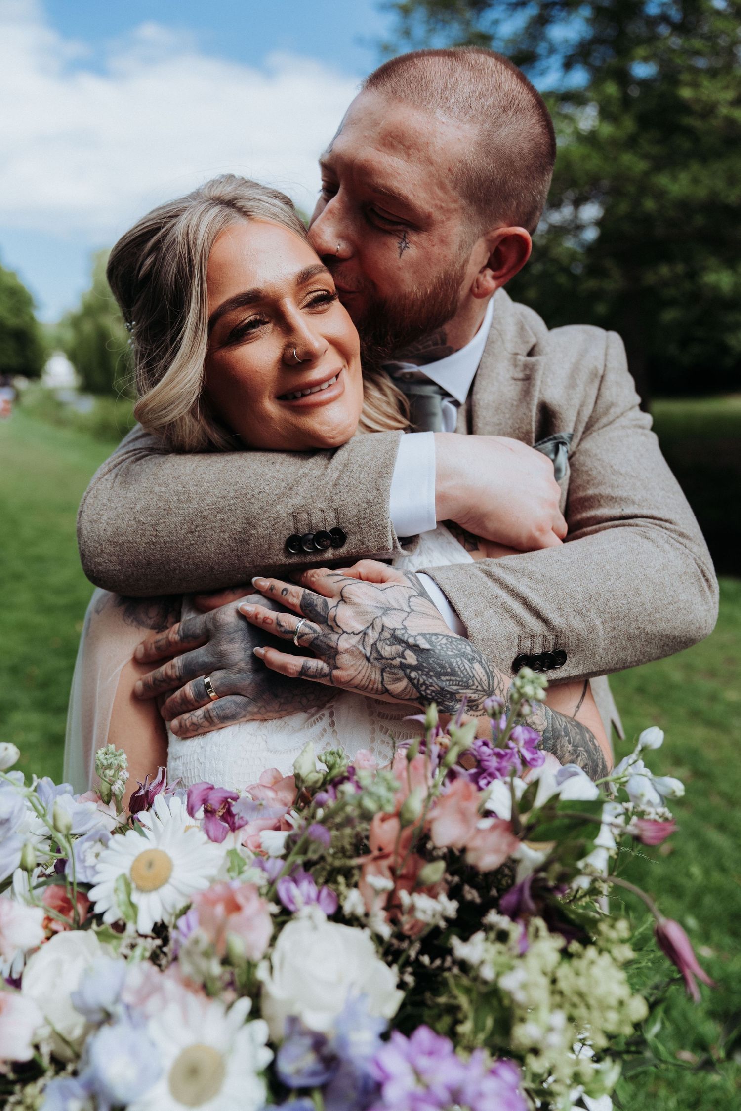 Emotional groom with tattoos embraces smiling bride from behind while she holds a colorful bridal bouquet.
