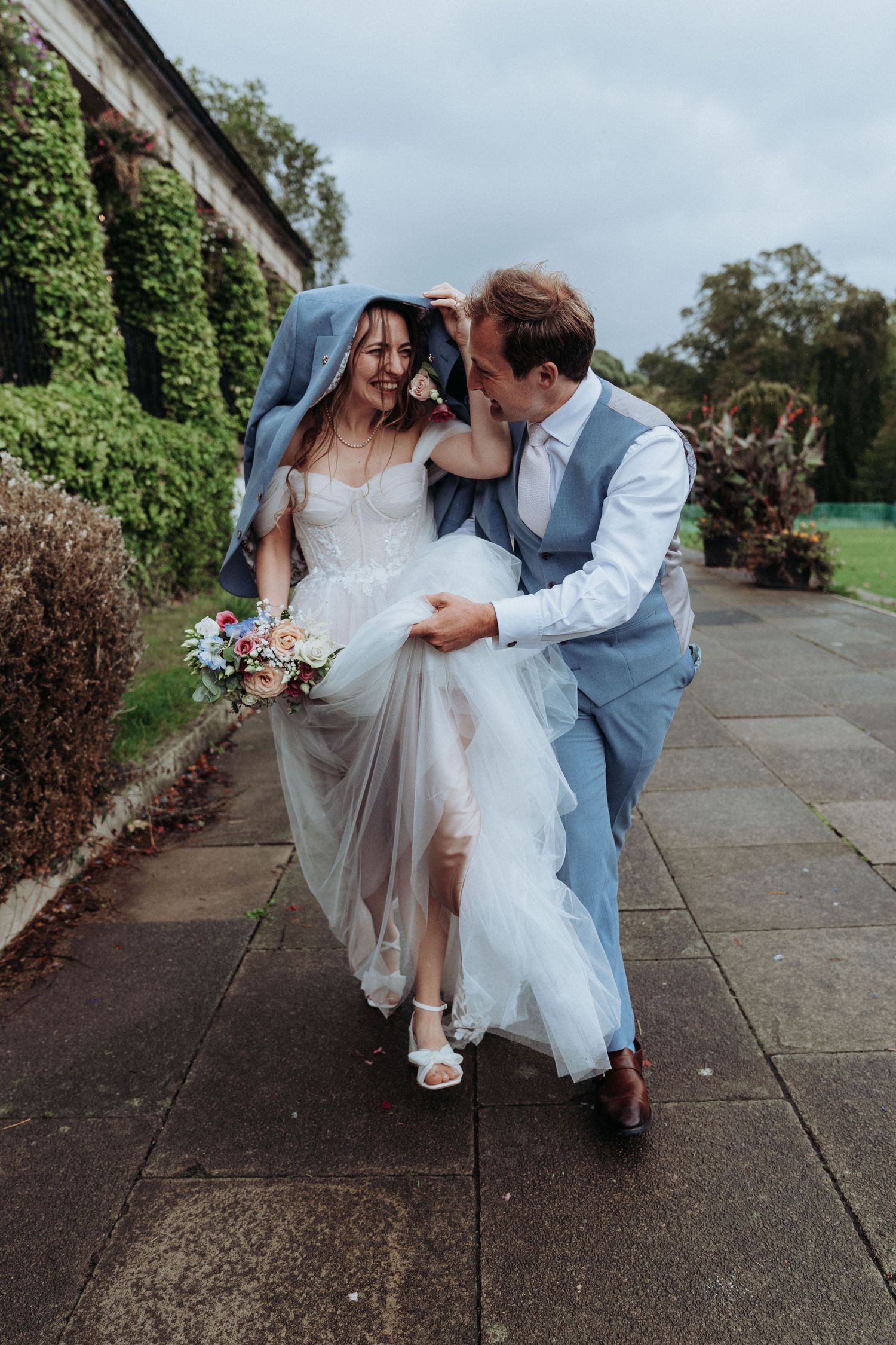Groom shelters laughing bride under his jacket while walking outdoors on a windy wedding day.