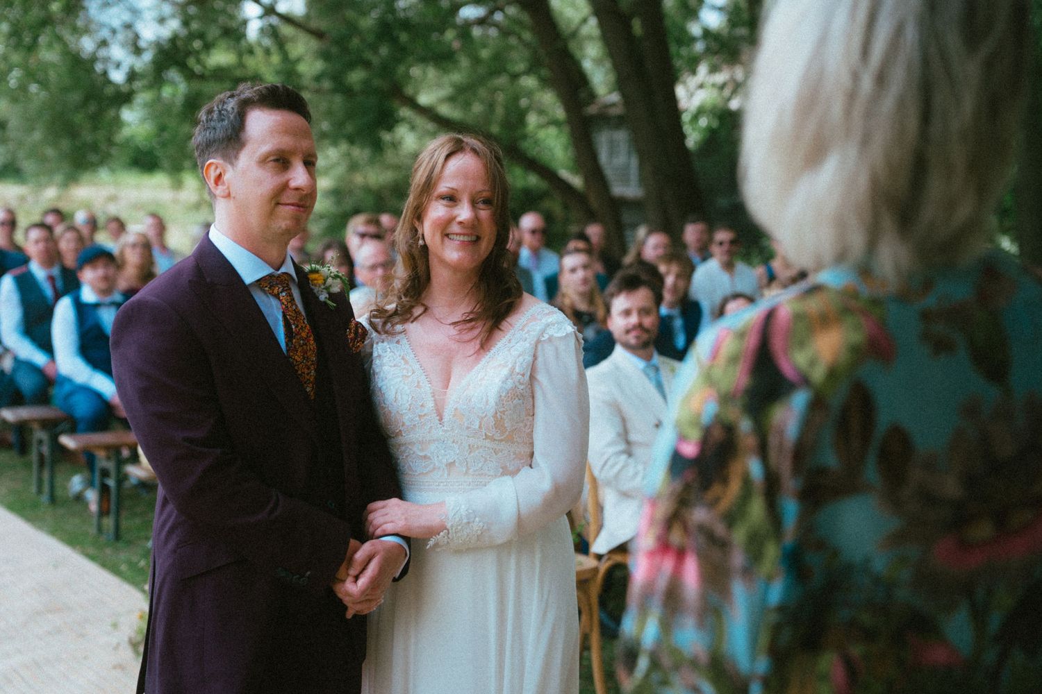 Bride and groom holding hands at outdoor wedding ceremony, smiling before officiant with guests seated in background.