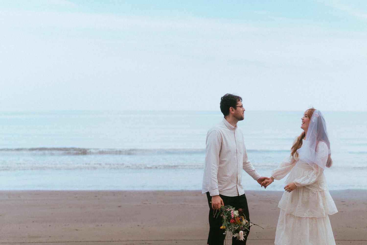 A bride and groom hold hands on a sandy beach, with the ocean in the background, on their wedding day.