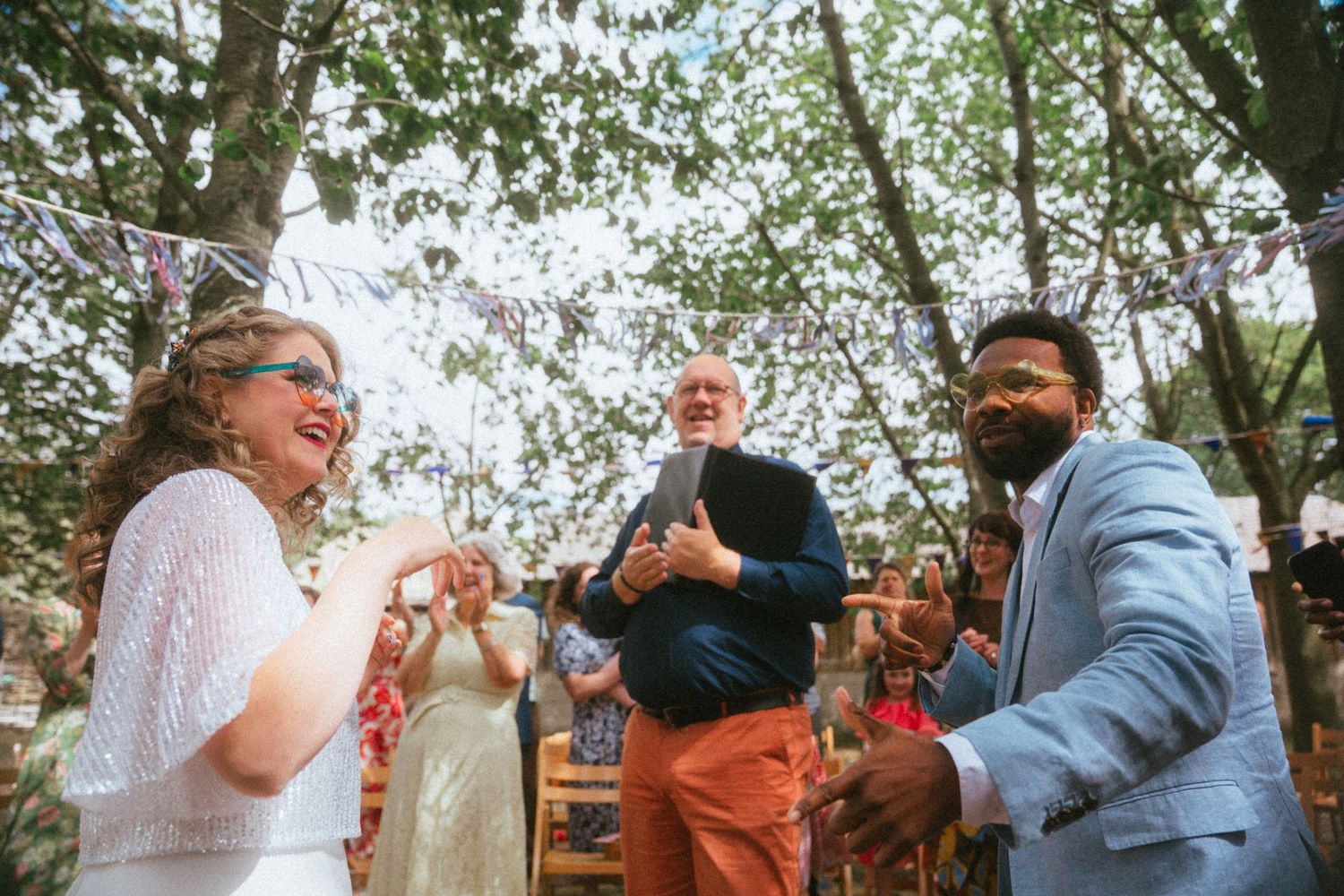 Joyful interracial couple laugh together during outdoor woodland wedding ceremony with colorful bunting.