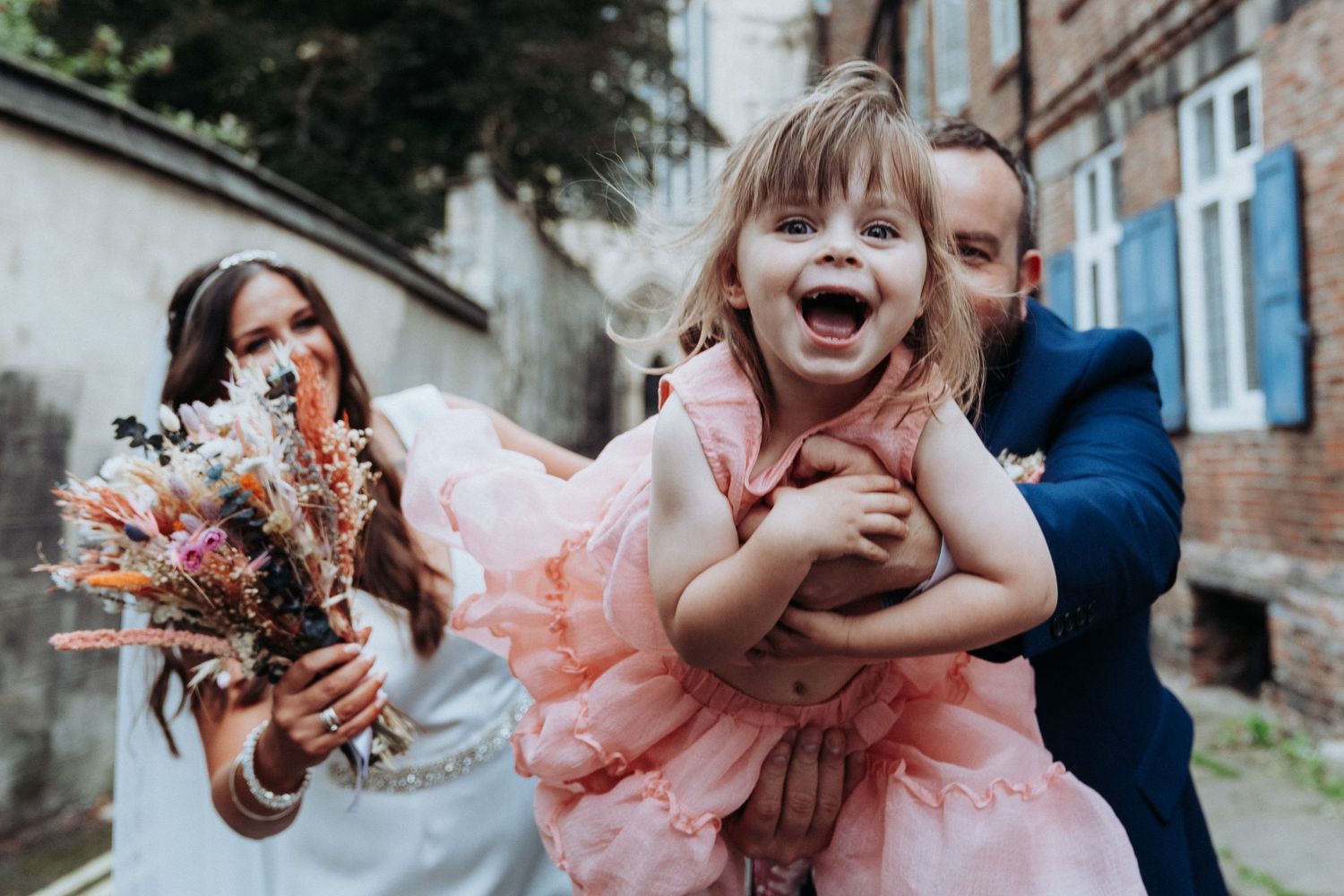 Beaming flower girl in pink tutu is lifted by bride and groom outside rustic brick building.