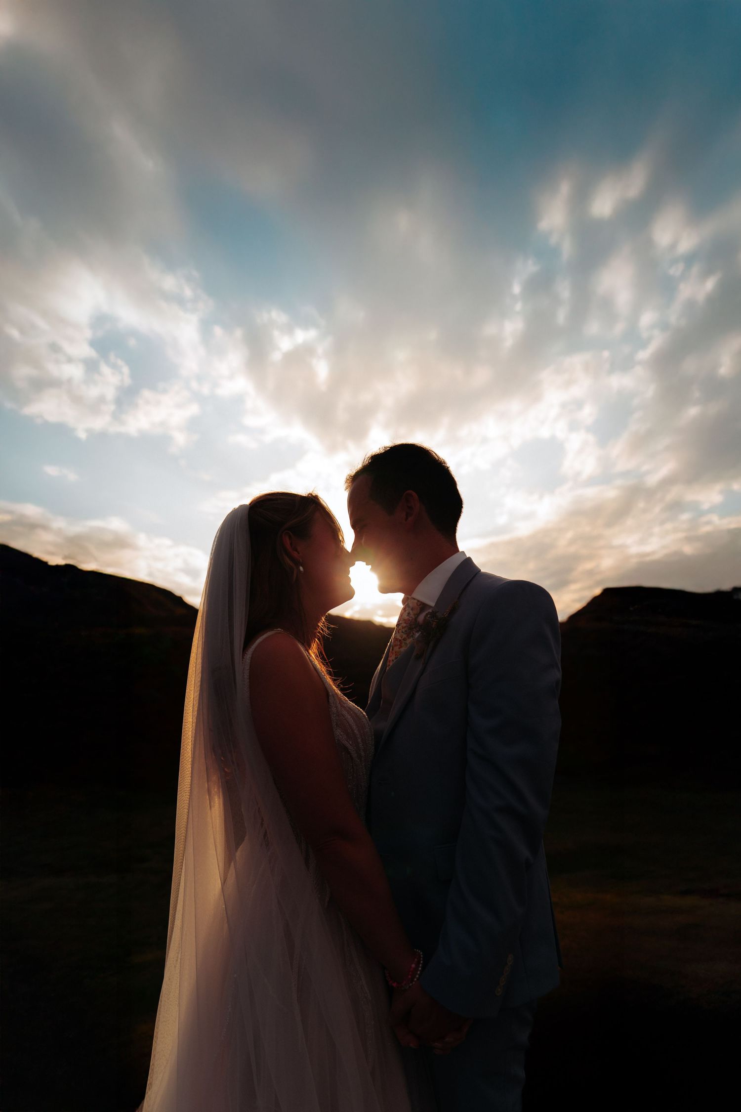 Silhouette of bride and groom holding hands at sunset with dramatic cloudy sky backdrop.