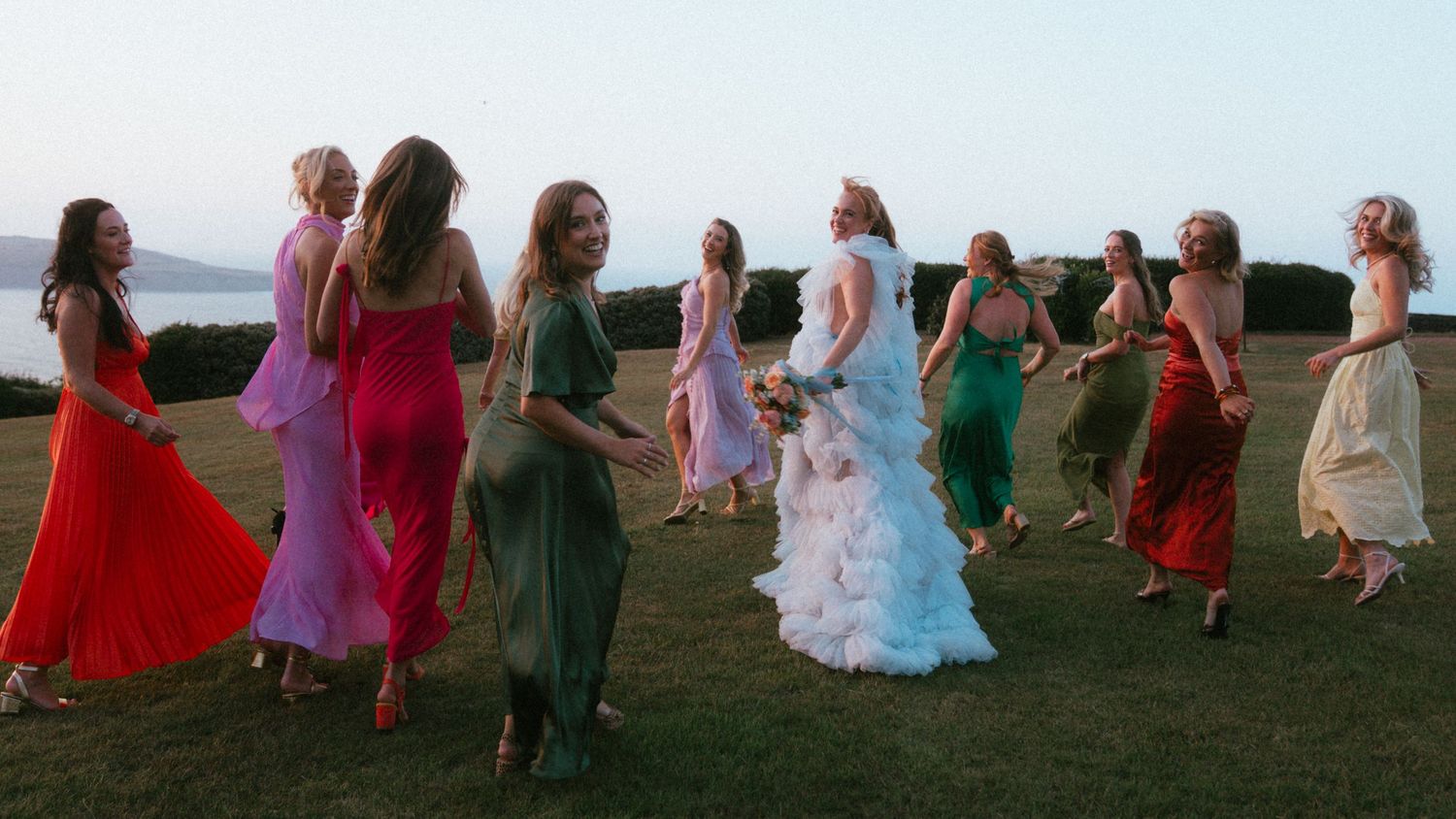 Bride in a white gown dances with bridesmaids in colorful dresses on a grassy lawn at a seaside wedding.