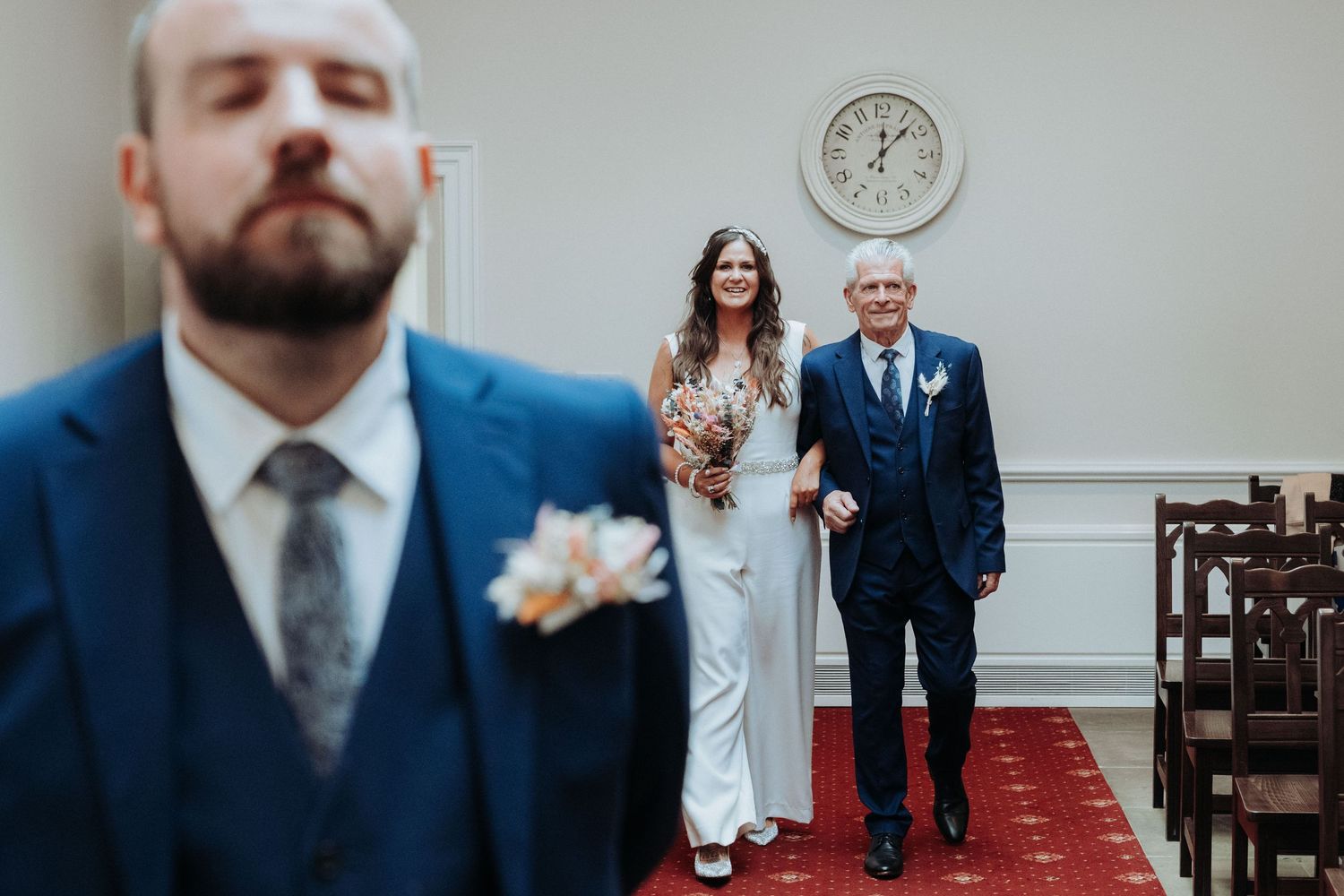 Bride walking down the aisle in white jumpsuit escorted by father toward waiting groom.
