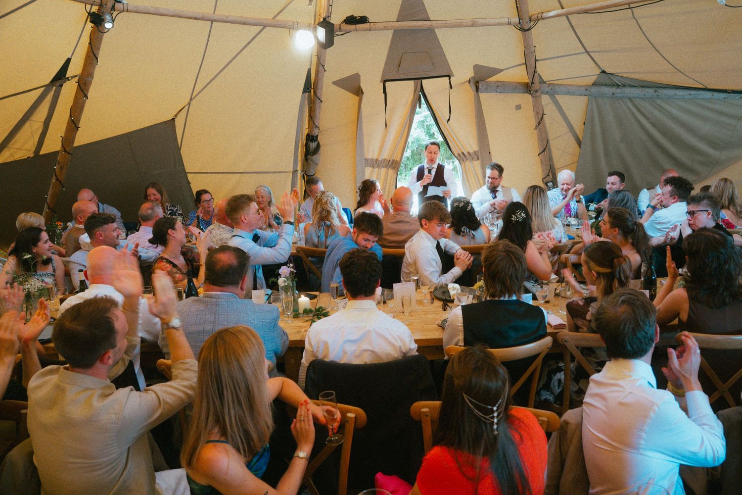 Wedding guests clap and celebrate during a speech inside a large decorated tipi reception tent.