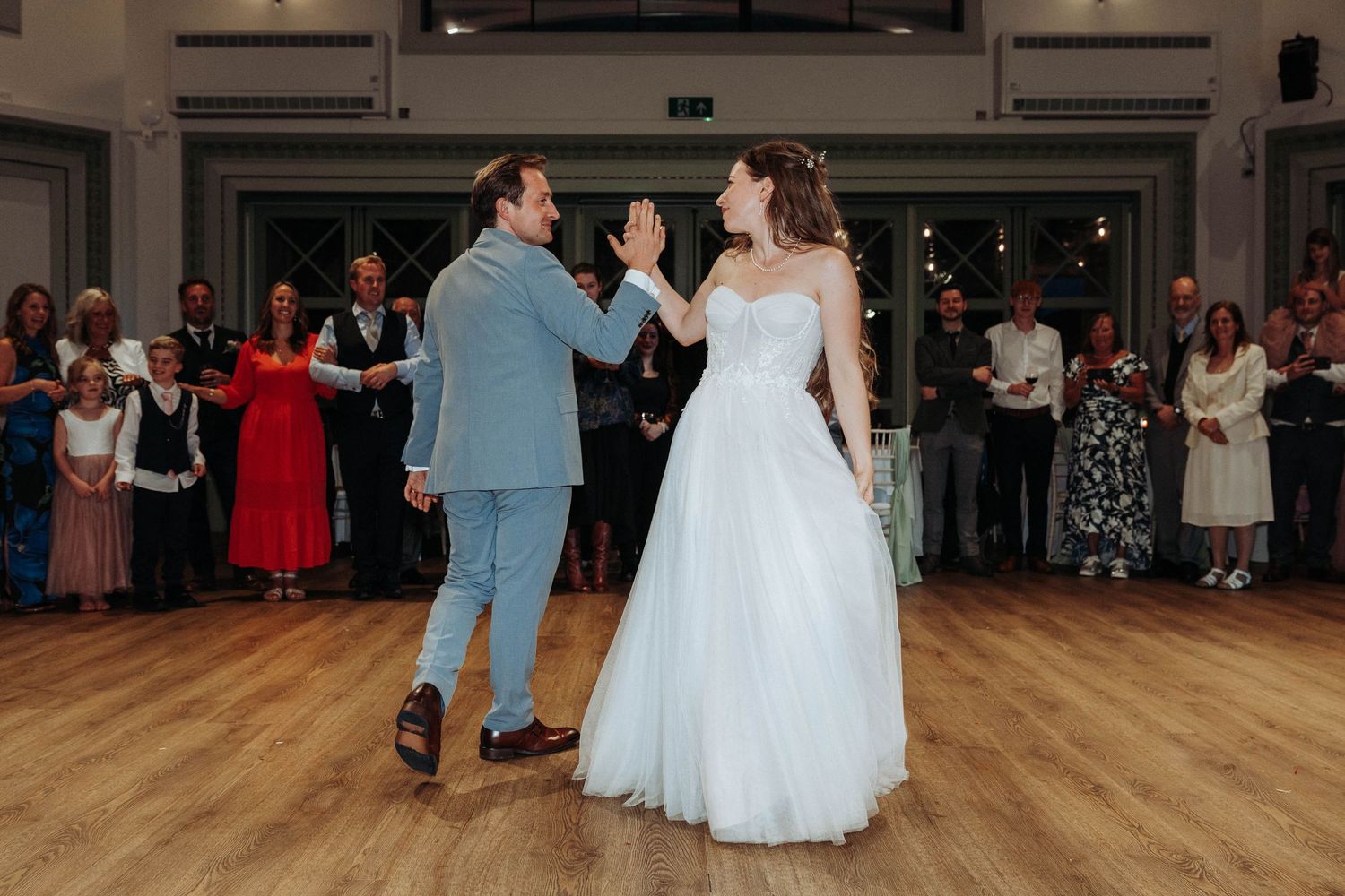 Bride in white ballgown and groom in blue suit sharing first dance before wedding guests.