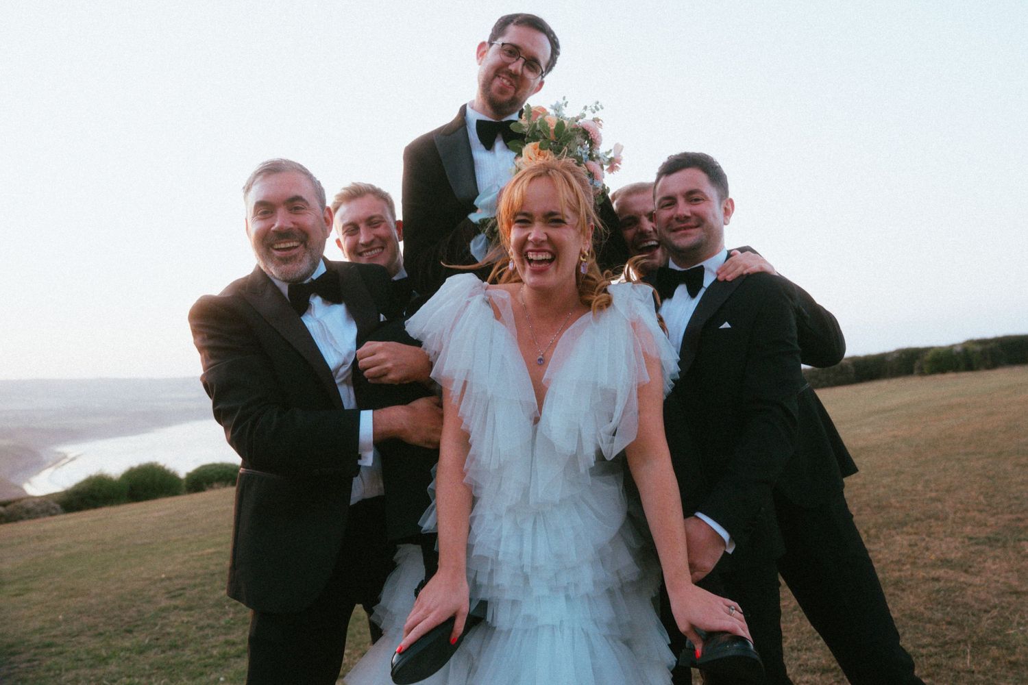 Laughing bride in a pale blue tulle dress poses with five groomsmen in black tuxedos on a grassy hill above the sea.