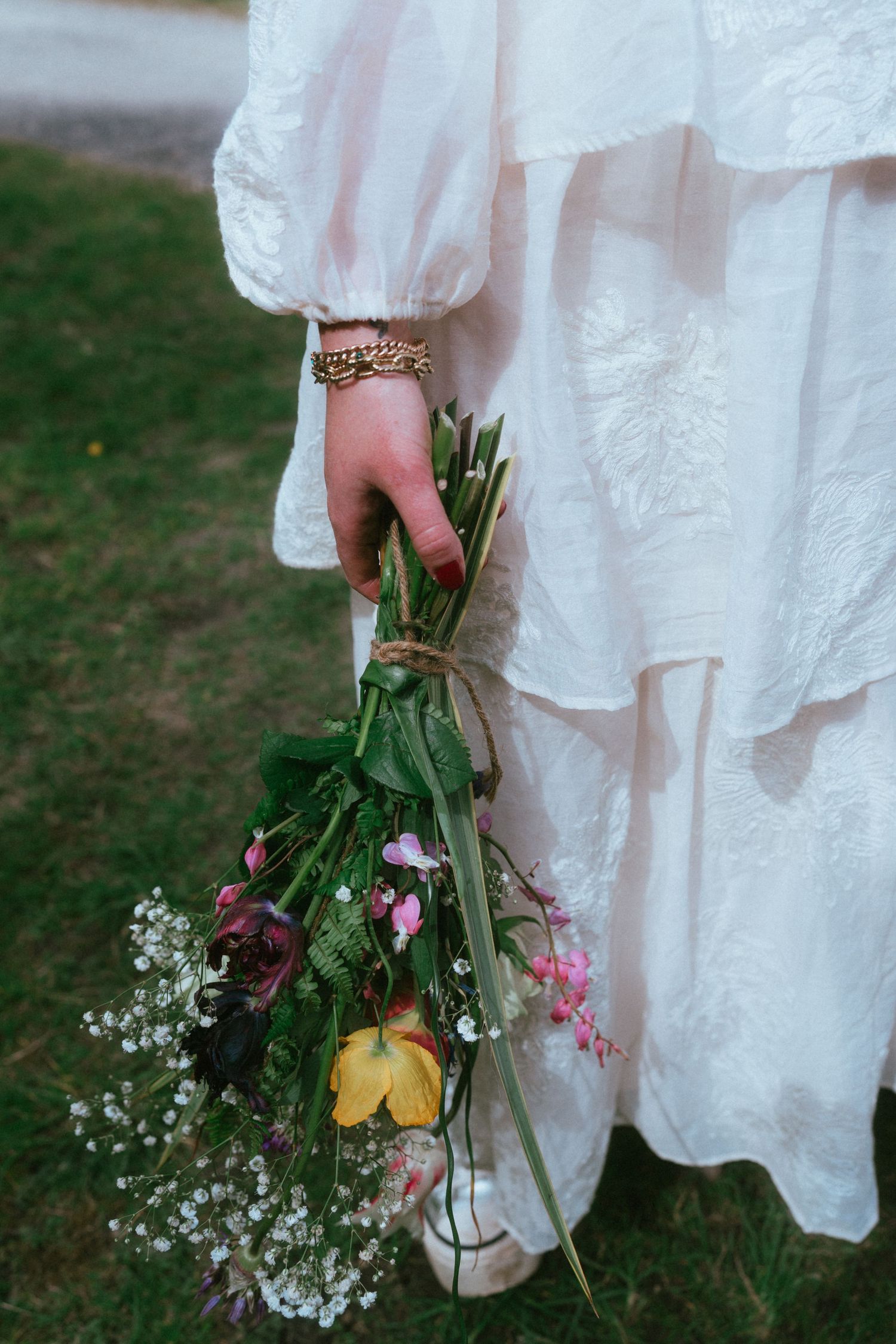 Woman in a white tiered embroidered dress holding a loose wildflower bouquet with baby's breath, tied with twine, on a green lawn.