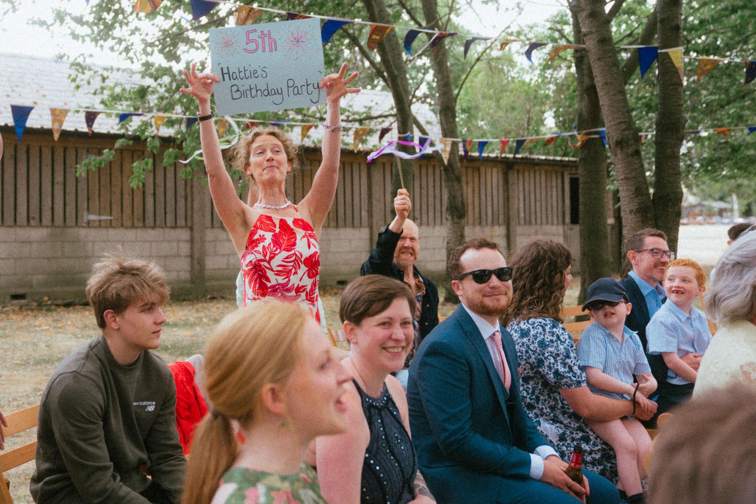 Guests celebrate at an outdoor birthday party with bunting decorations and a woman holding a sign.