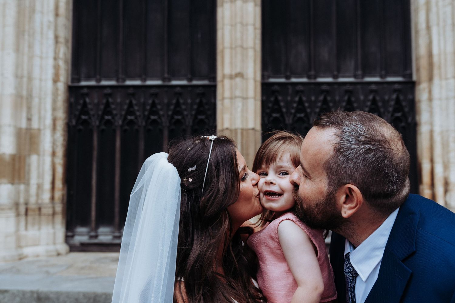 Bride and groom simultaneously kiss smiling flower girl in pink dress on cheeks outside cathedral.