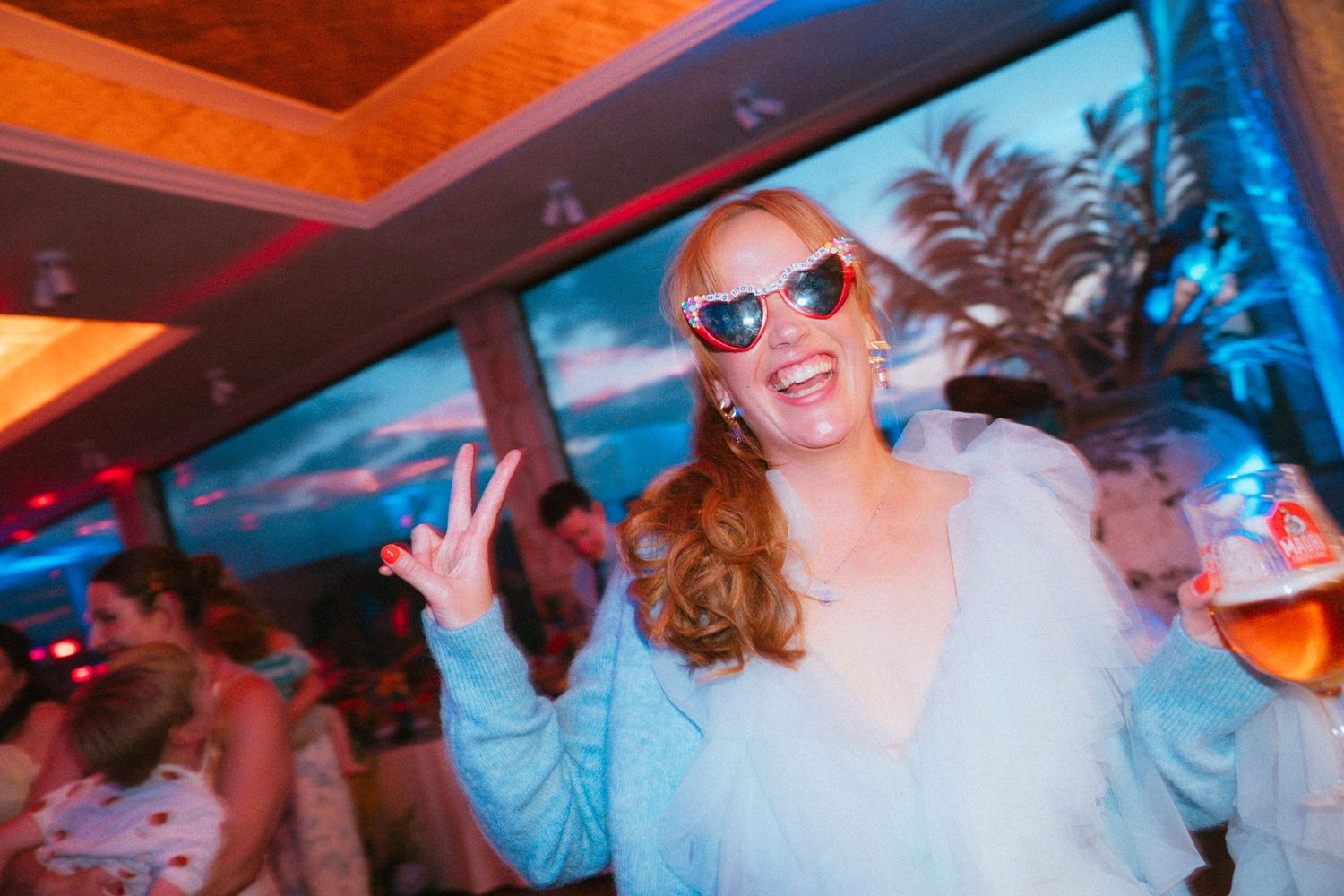Smiling woman in heart-shaped sunglasses and a light blue fluffy jacket flashes a peace sign while holding a drink at a lively party.