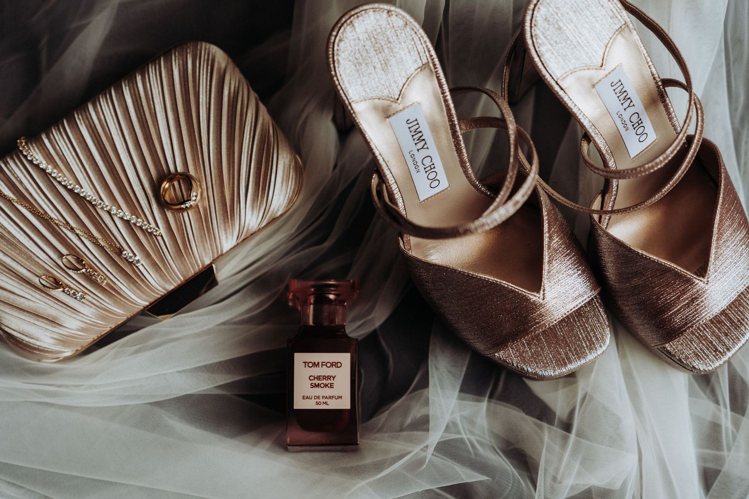 Flat lay of Jimmy Choo wedding shoes, Tom Ford perfume, and a pleated clutch bag.