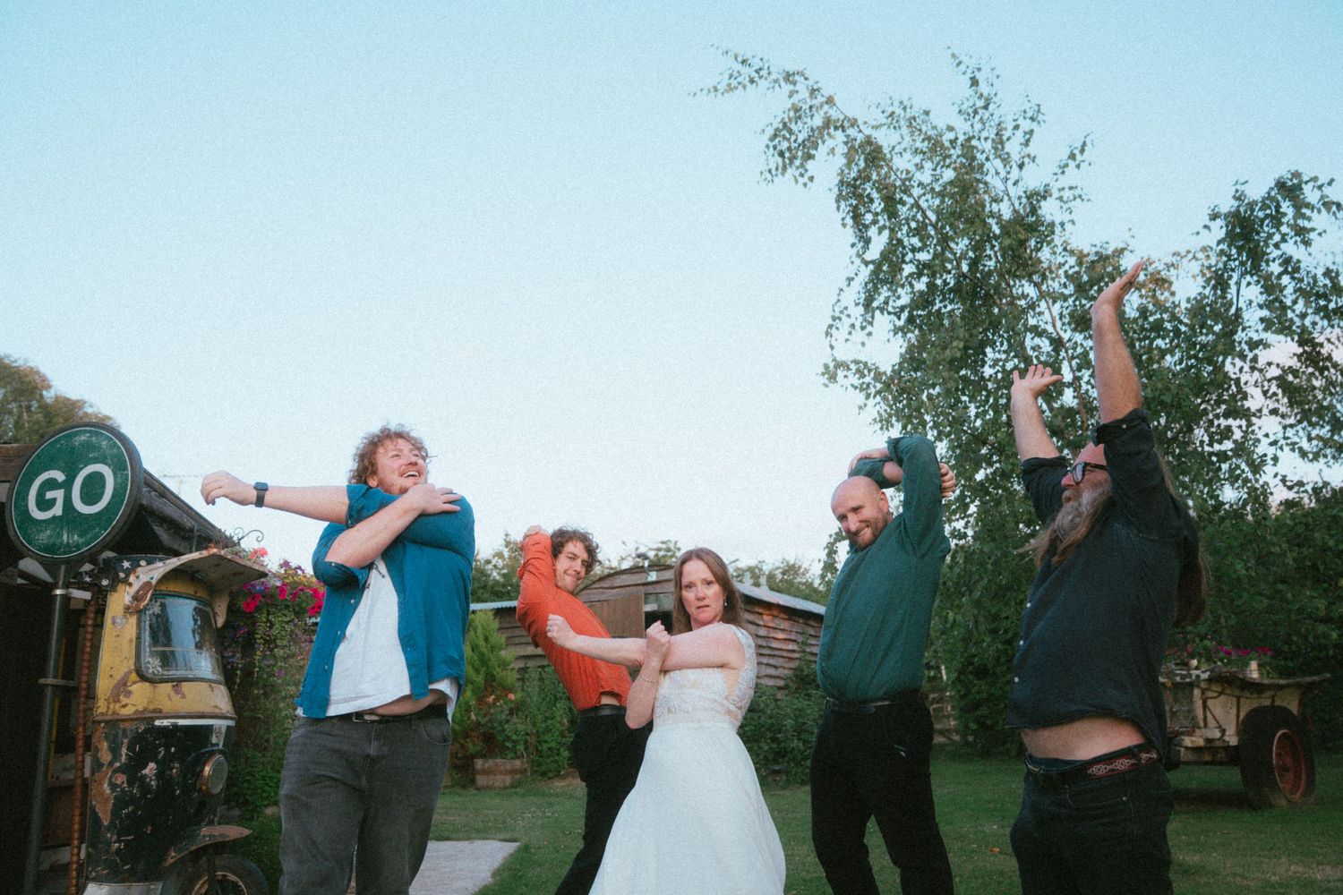 Bride in white gown celebrating outdoors with wedding guests cheering and dancing near rustic venue.