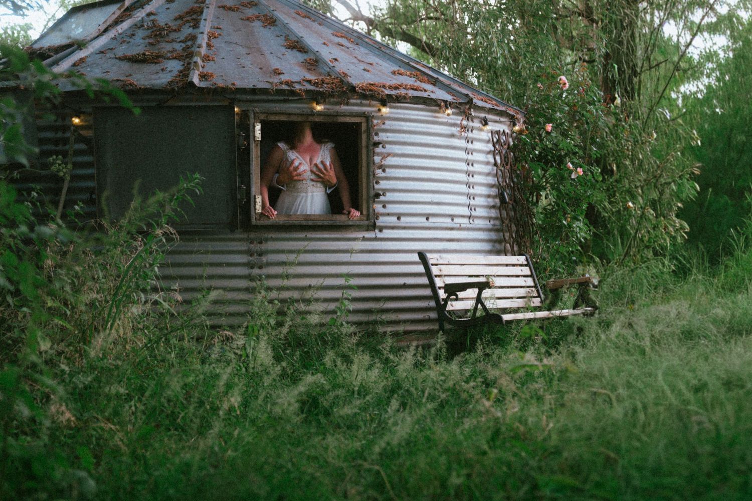 Bride in white dress peers through the window of a rustic corrugated metal garden building.