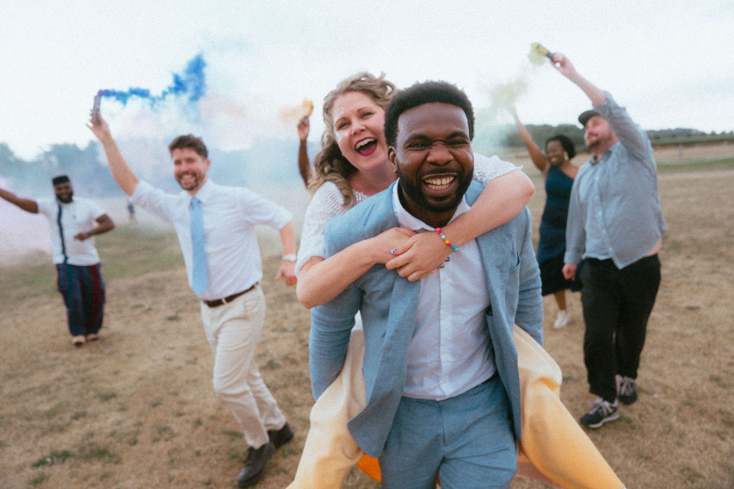 Joyful interracial wedding couple with guests celebrating in a field with colorful blue and yellow smoke bombs.