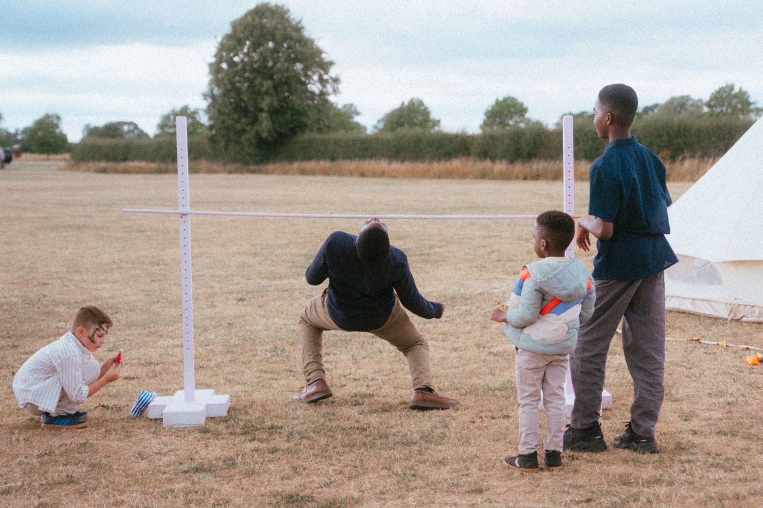 Adults and children playing a lawn game with white poles in an open field on a cloudy day.