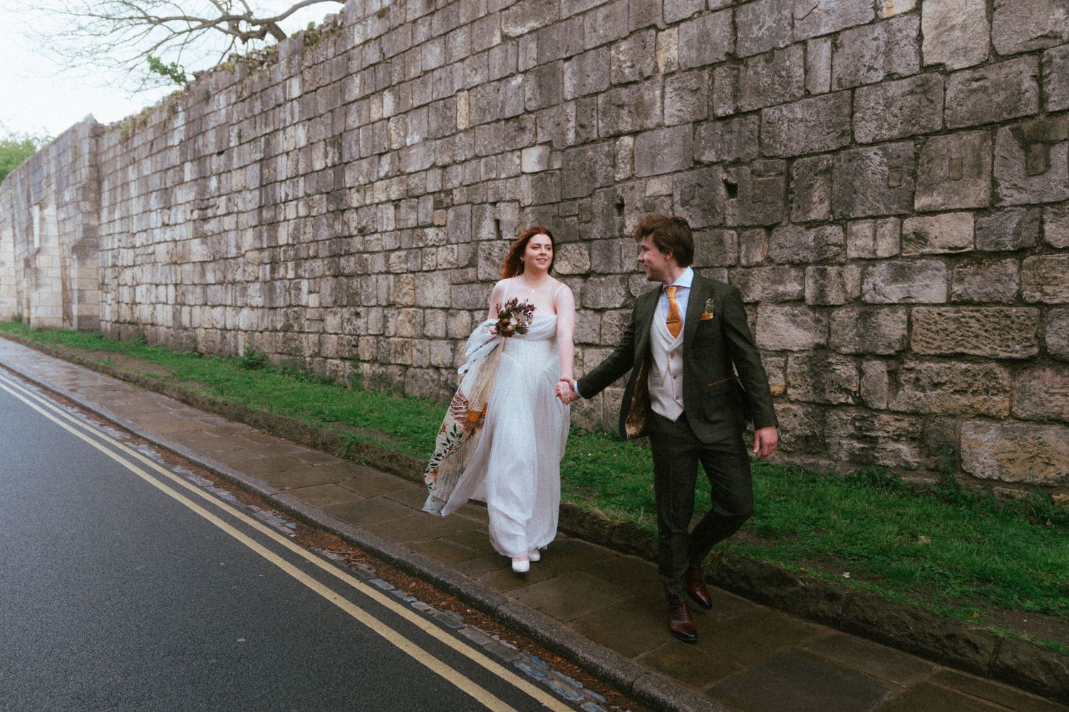 Bride and groom walk along cobblestone street beside historic stone wall, bride wearing floral sleeve wedding dress.