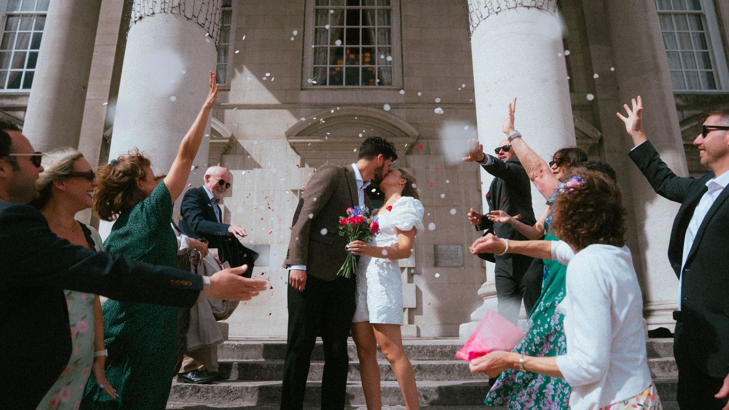 Bride and groom celebrated with confetti outside a grand building surrounded by smiling wedding guests.