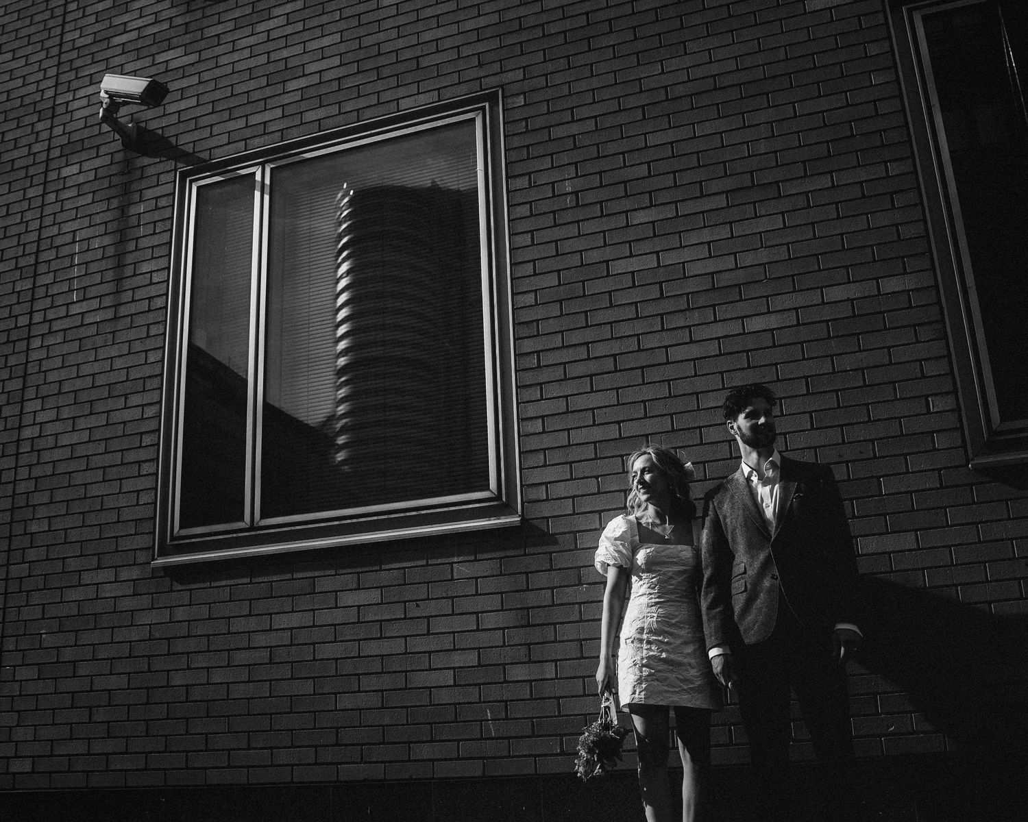 Black and white photo of couple standing together against a brick wall beneath a large window.