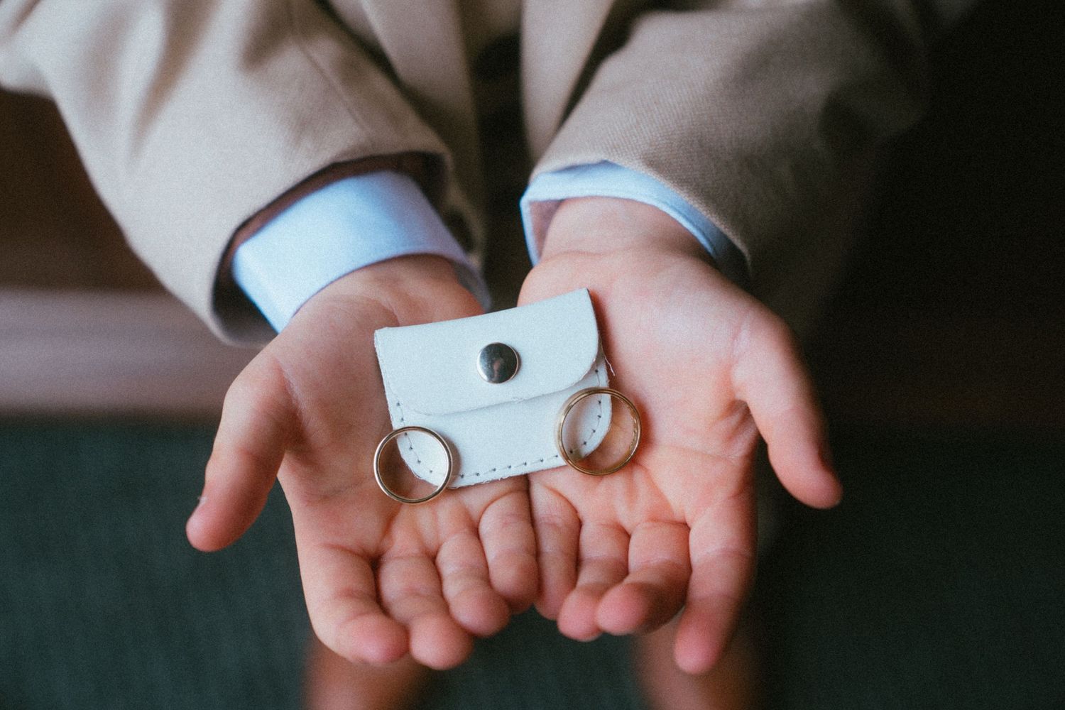 Groom holding two gold wedding rings and a small white ring pouch in his outstretched hands.