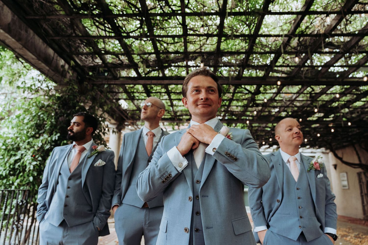 Groom in light blue suit adjusting tie with groomsmen under vine-covered pergola outdoors.