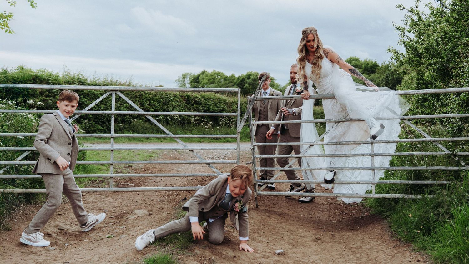 Candid wedding photo of bride and groom with children by a farm gate, with one boy falling in the mud.