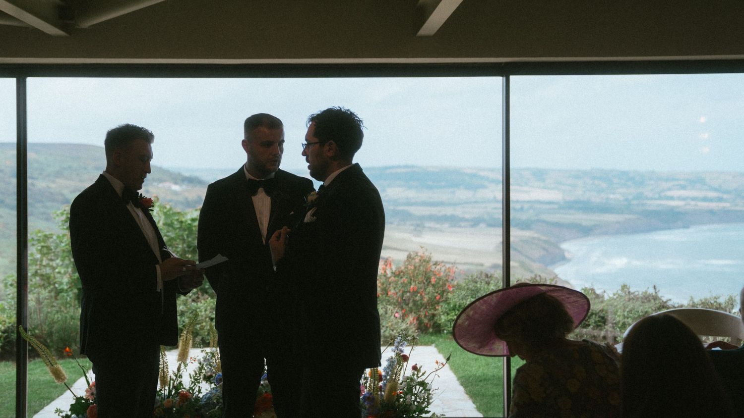 Three men in dark suits stand together during a wedding ceremony in a glass-walled venue overlooking a scenic coastline and sea.