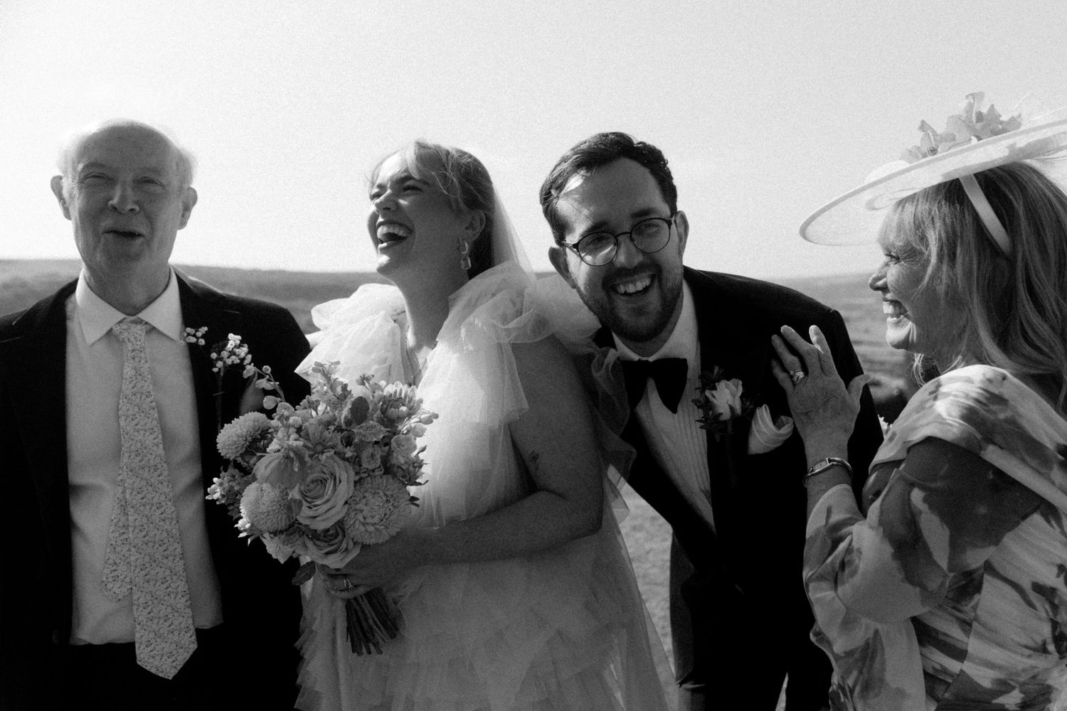 Black and white photo of a laughing bride in a ruffled gown holding a bouquet, surrounded by joyful wedding guests in formal attire.