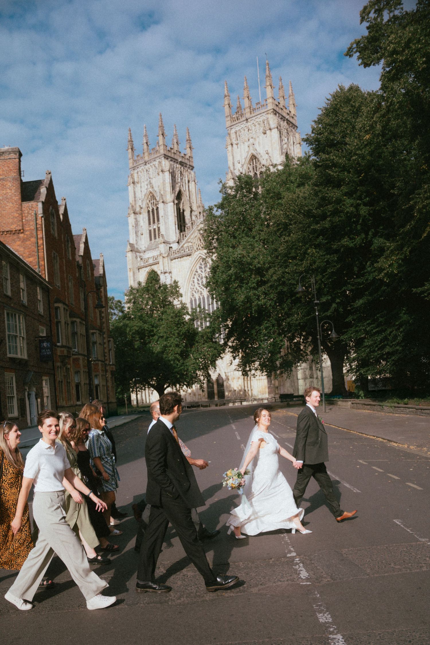Bride in a white gown and wedding guests crossing a city street with a Gothic cathedral and tall green trees in the background.